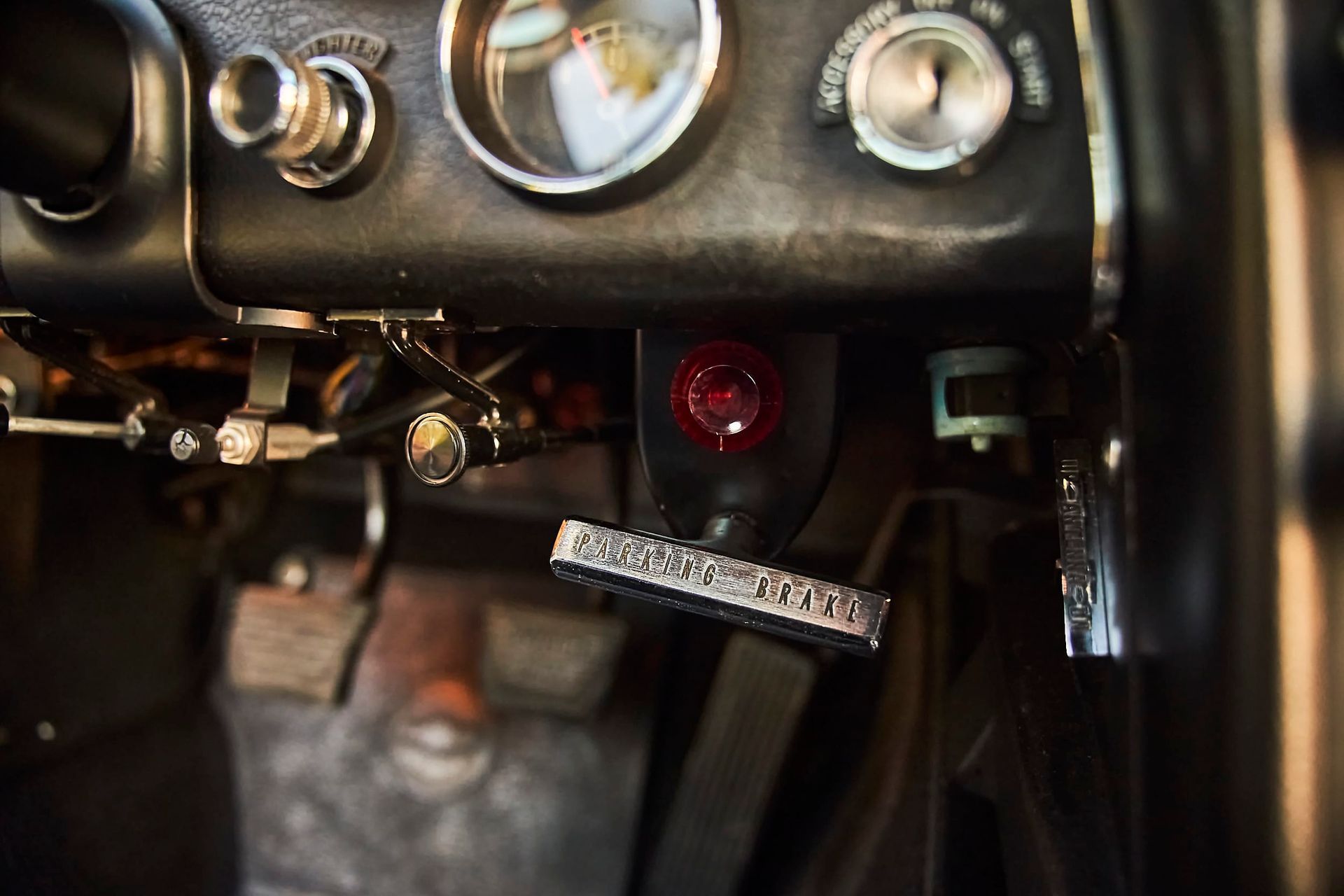 Close-up of a vintage car's dashboard. Visible are the pedals, a red warning light, and various dials.