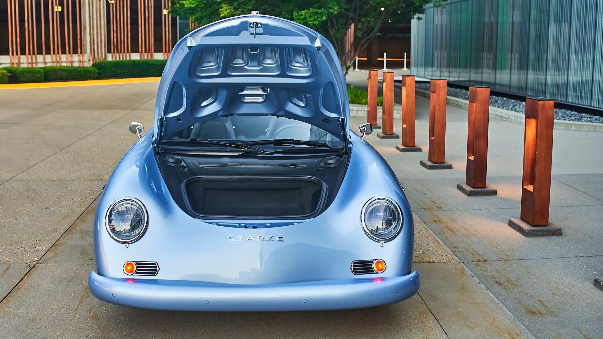 Light blue classic Porsche 356, hood open, parked on pavement, near modern architecture with wooden posts.