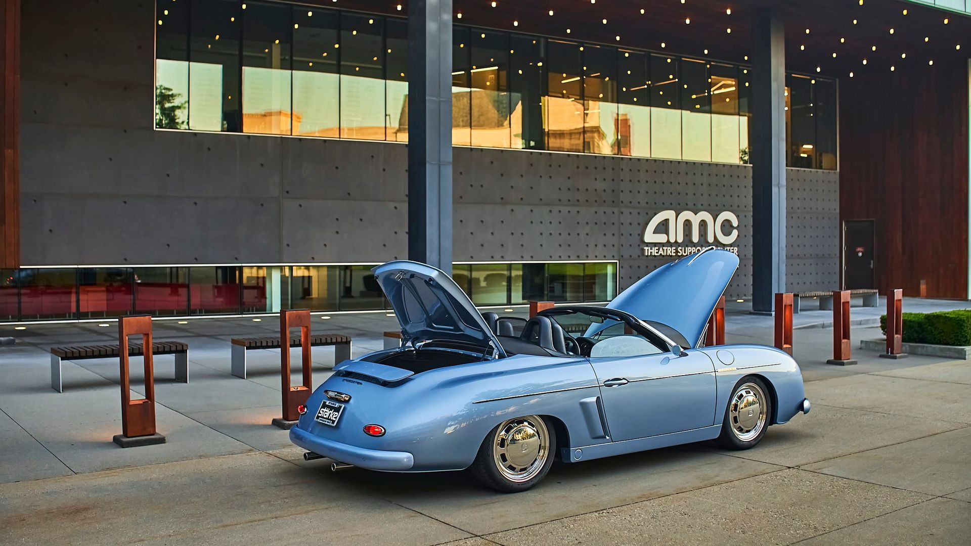 A light blue classic convertible with open hoods parked in front of an AMC theater. The setting is urban, with a modern building in the background.