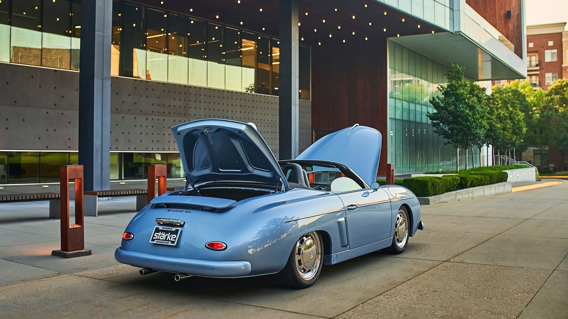 Blue classic convertible with open hood parked in front of a modern building.