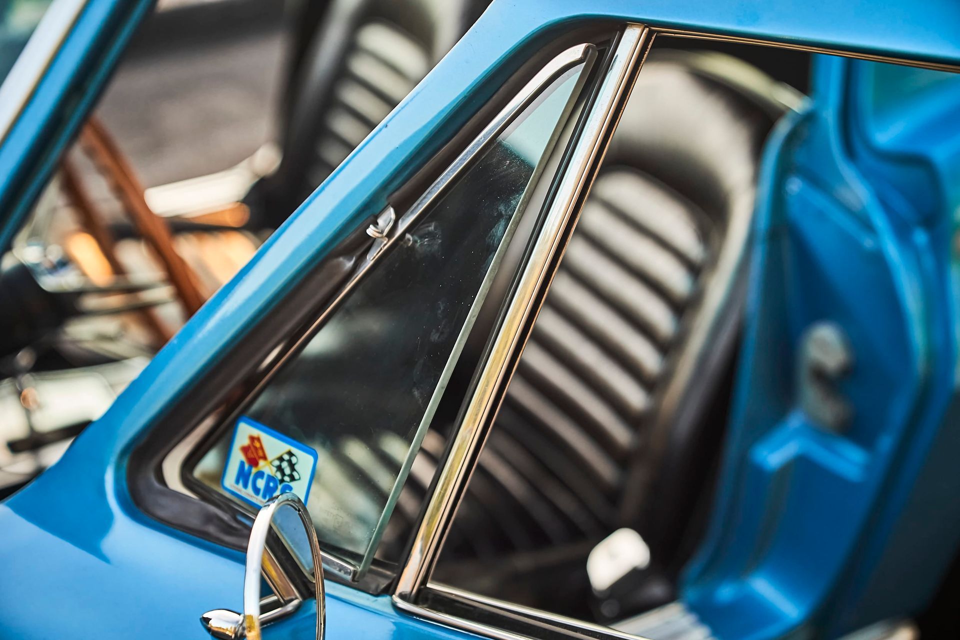 Close-up of a blue classic car's open window with black interior visible. A sticker is affixed to the glass.
