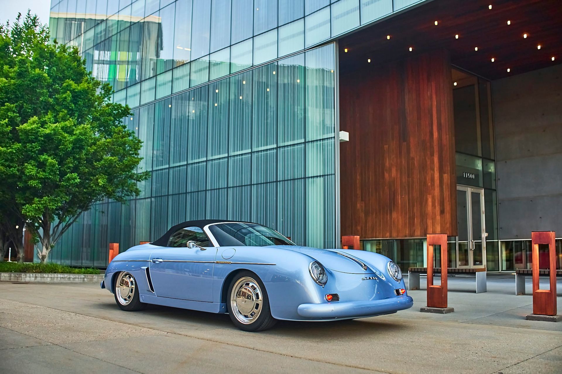 Light blue vintage convertible Porsche parked outside a modern building with glass and wood paneling.