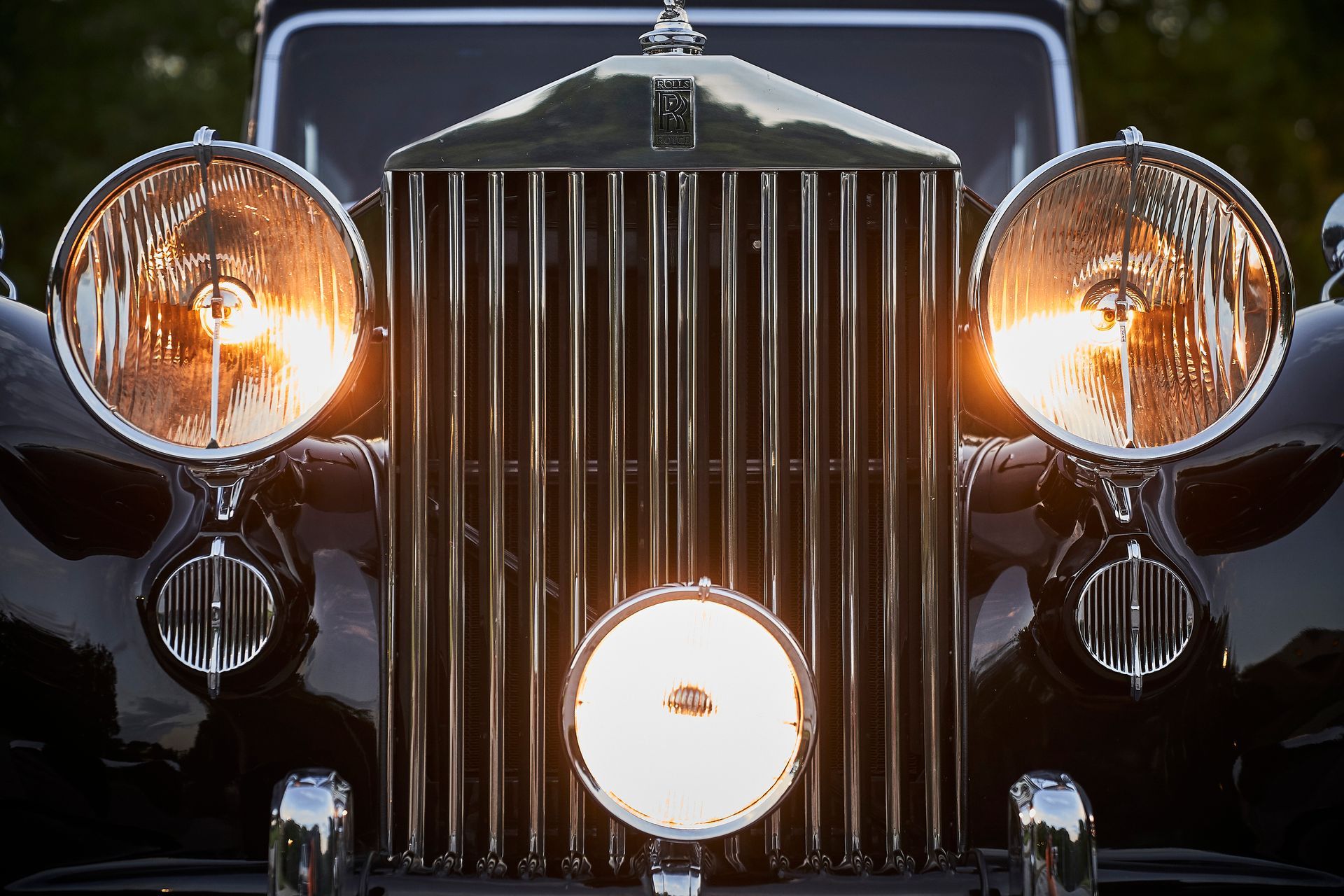 The front of an old rolls royce car with its lights on.