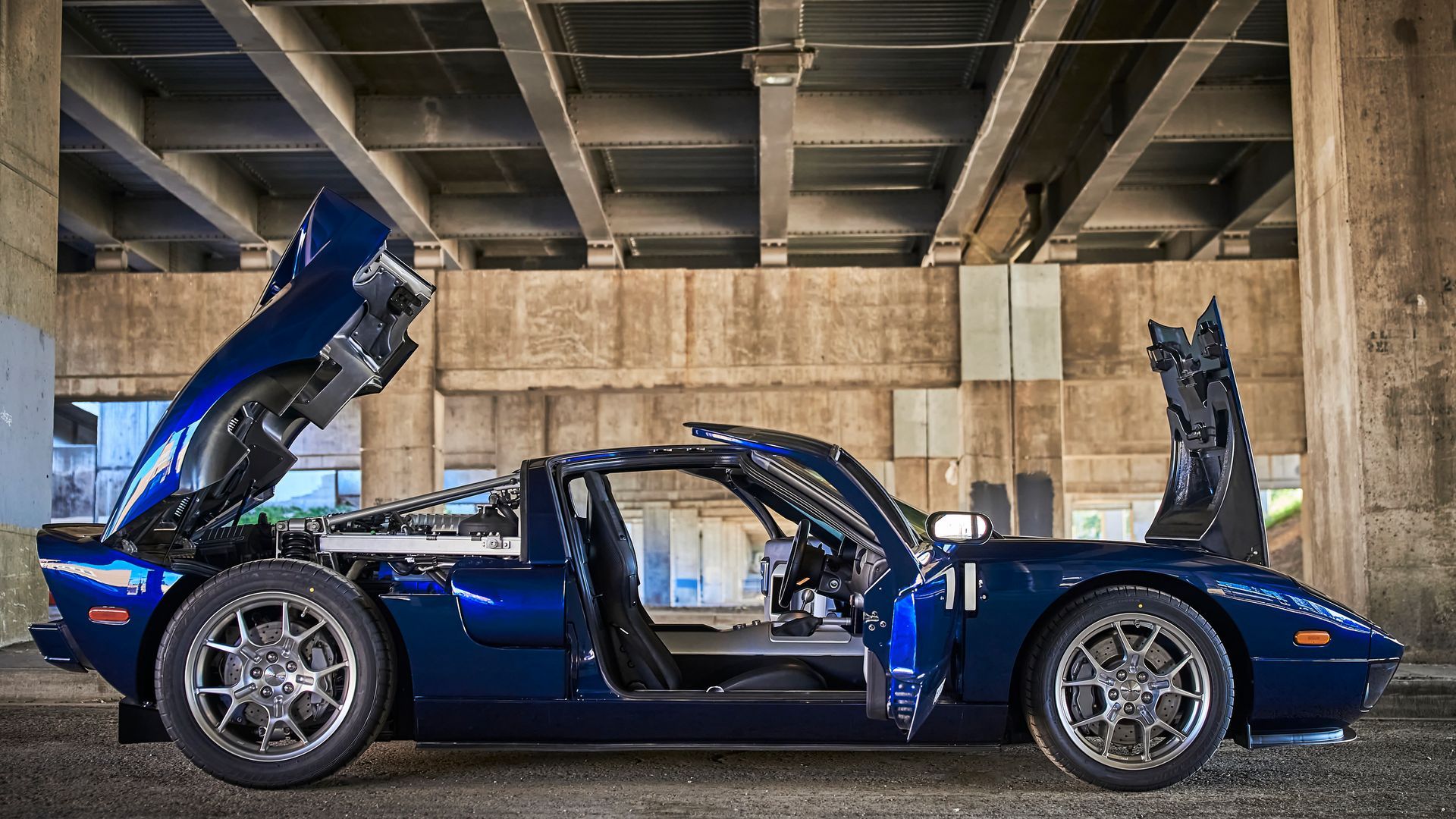 A blue sports car with its hood up is parked under a bridge.