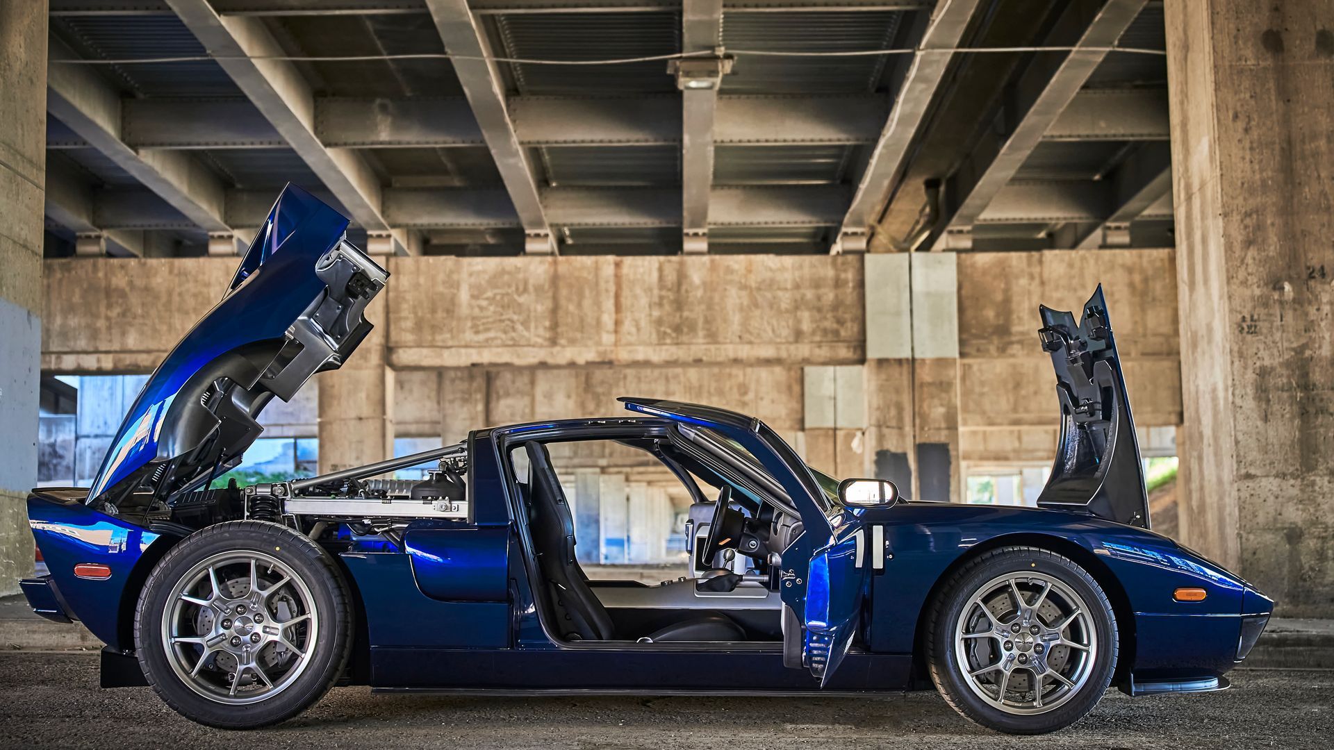 A blue sports car with its hood open is parked under a bridge.
