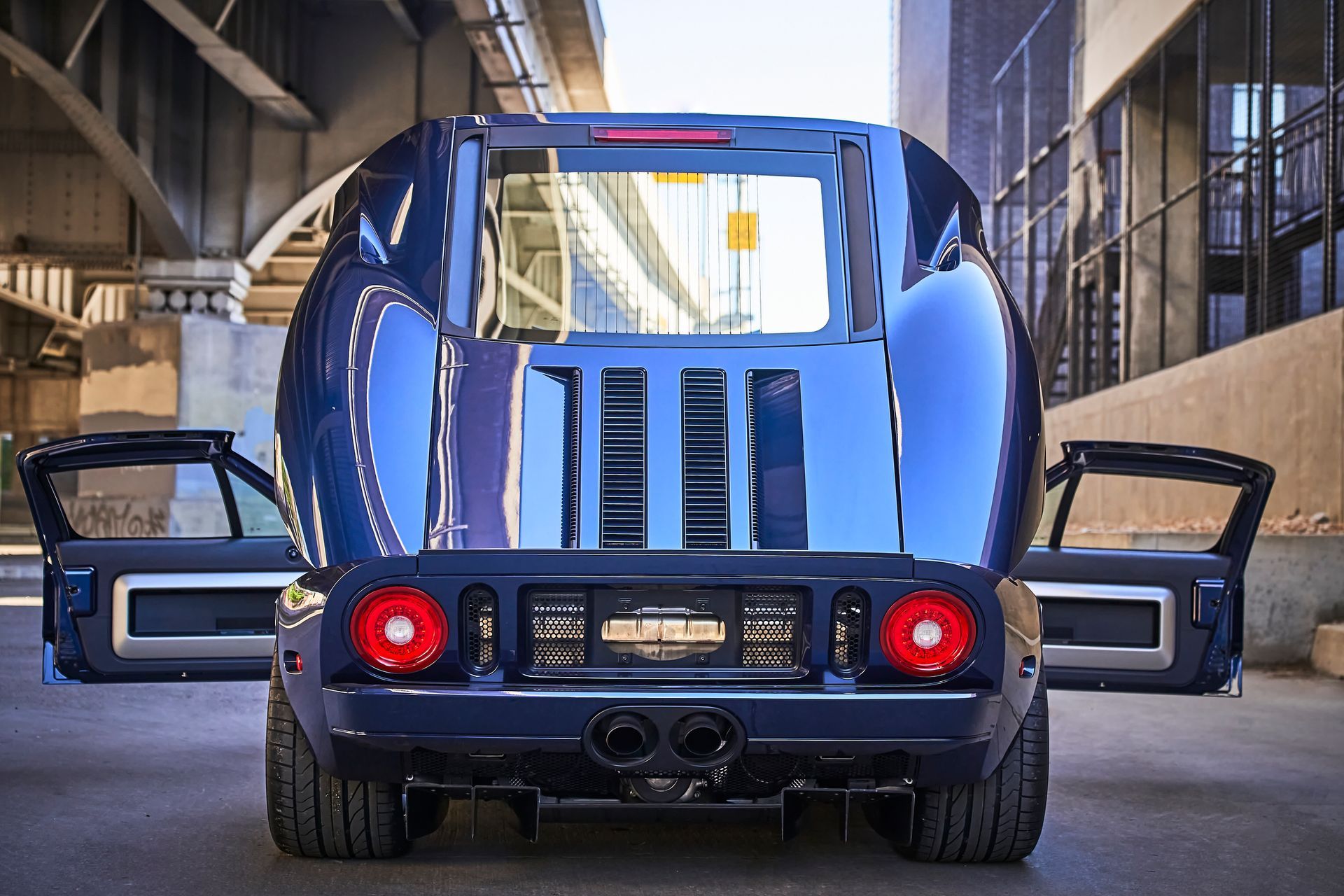 A blue sports car with its doors open is parked under a bridge.