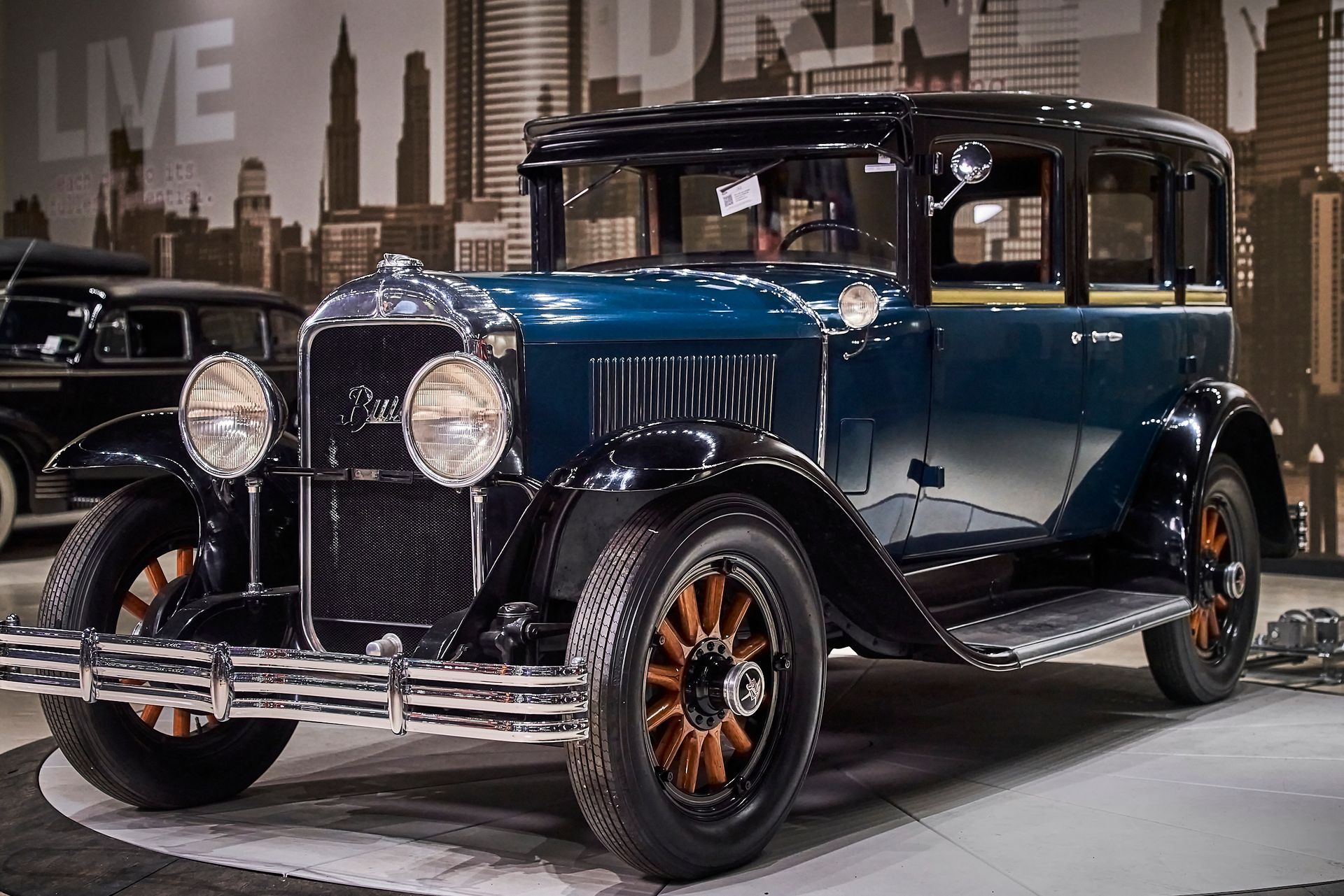 A blue and black vintage car is parked in front of a city skyline.