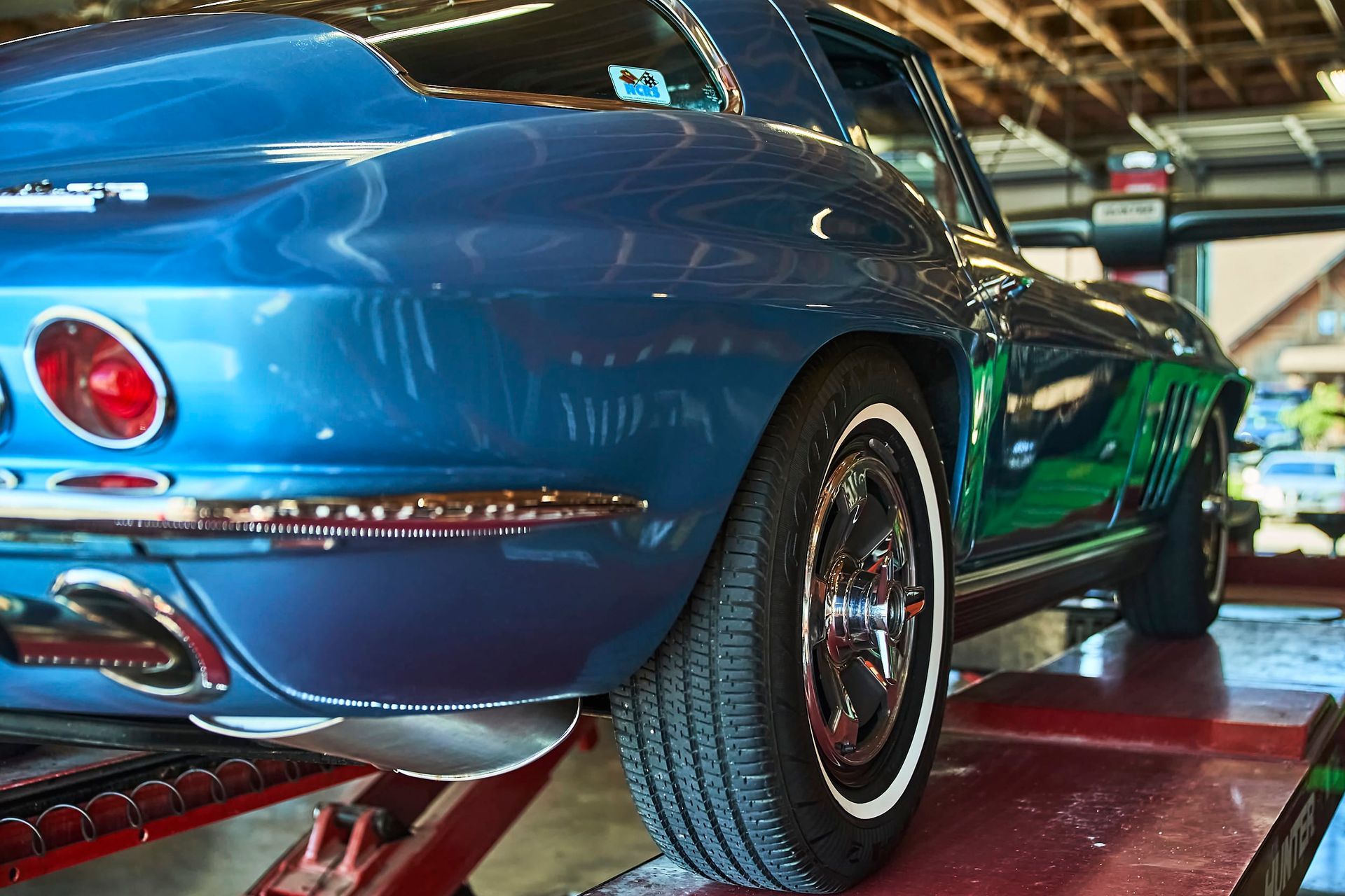 Blue classic Corvette on a lift, rear view. Features white-wall tires, chrome accents, and is in a garage setting.
