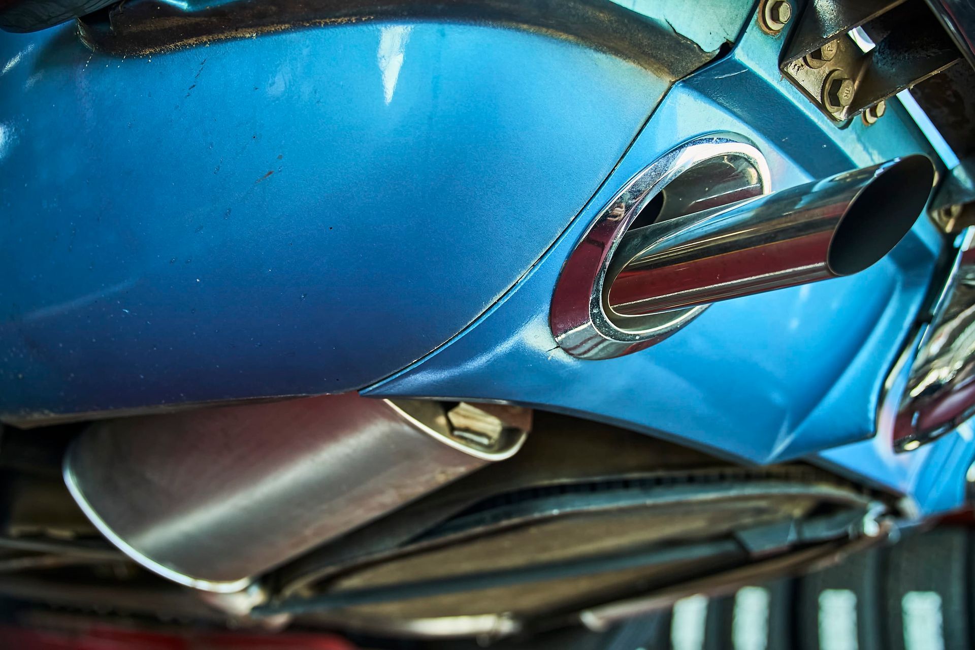 Blue car's exhaust pipes, chrome-trimmed, under a blue painted panel. Bottom view, with a shiny metal muffler visible.