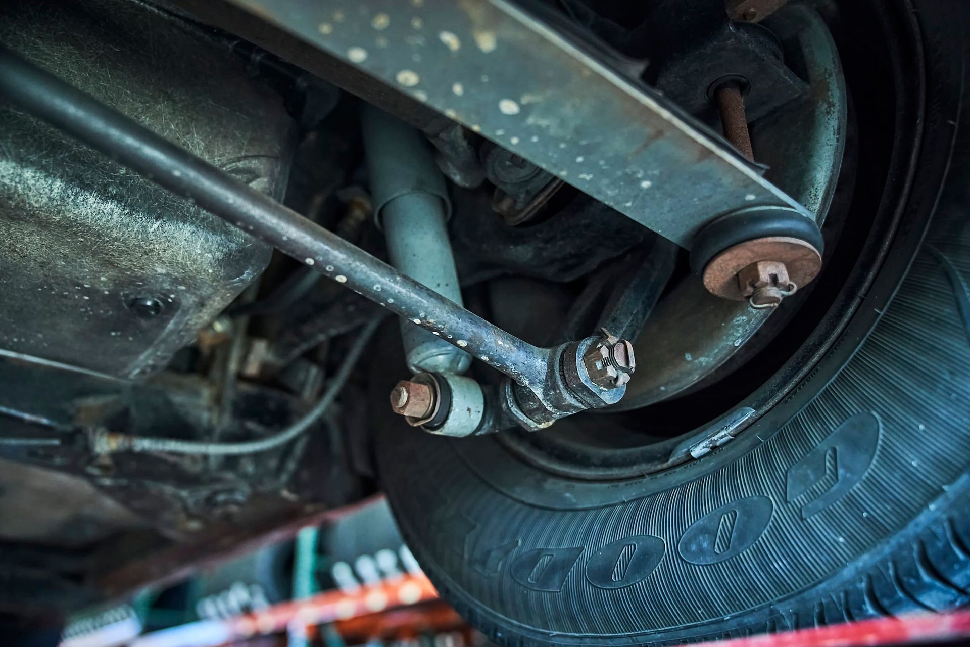 Close-up view of a car's suspension system, including the shock absorber, control arm, and tire. The components are metal and have some surface rust.