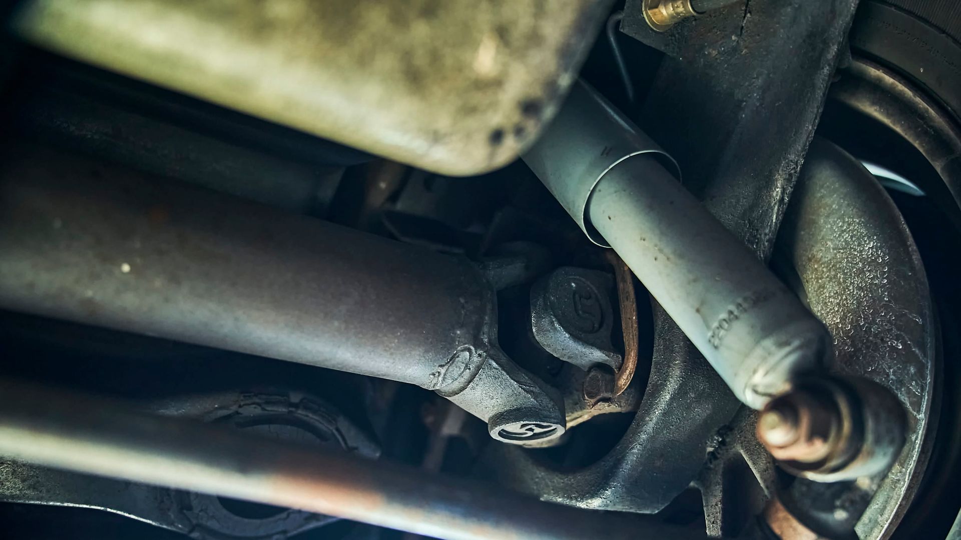 Undercarriage view showing a rusted driveshaft and shock absorber. The setting appears to be a vehicle repair shop.