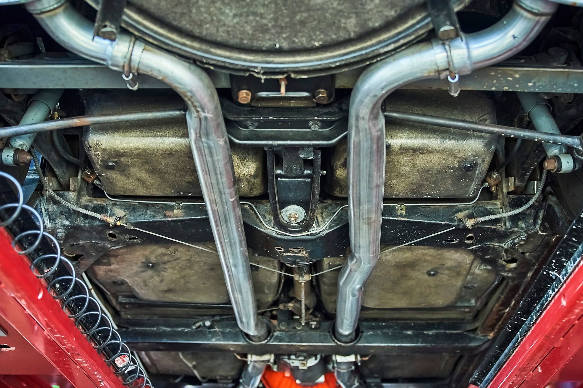 Underside view of a classic car, showing the exhaust system and undercarriage. The car is red, with silver pipes.