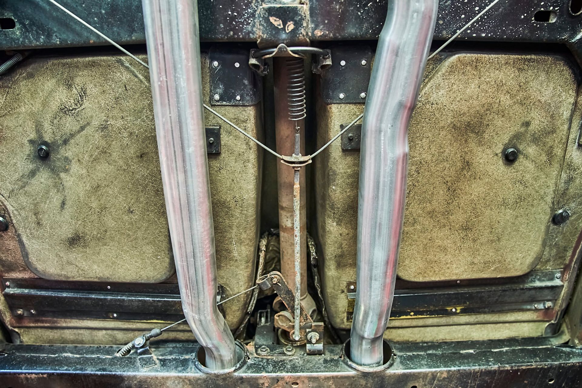 Underside view of a car, showing the exhaust pipes, driveshaft, and undercarriage insulation. The parts are rusty and dirty.