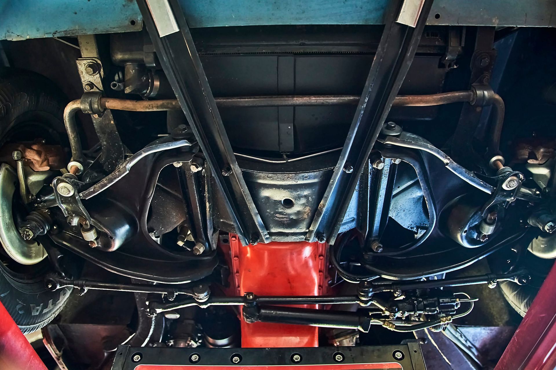 Underside view of a classic car chassis in a garage, showcasing the suspension and undercarriage components, painted black and red.