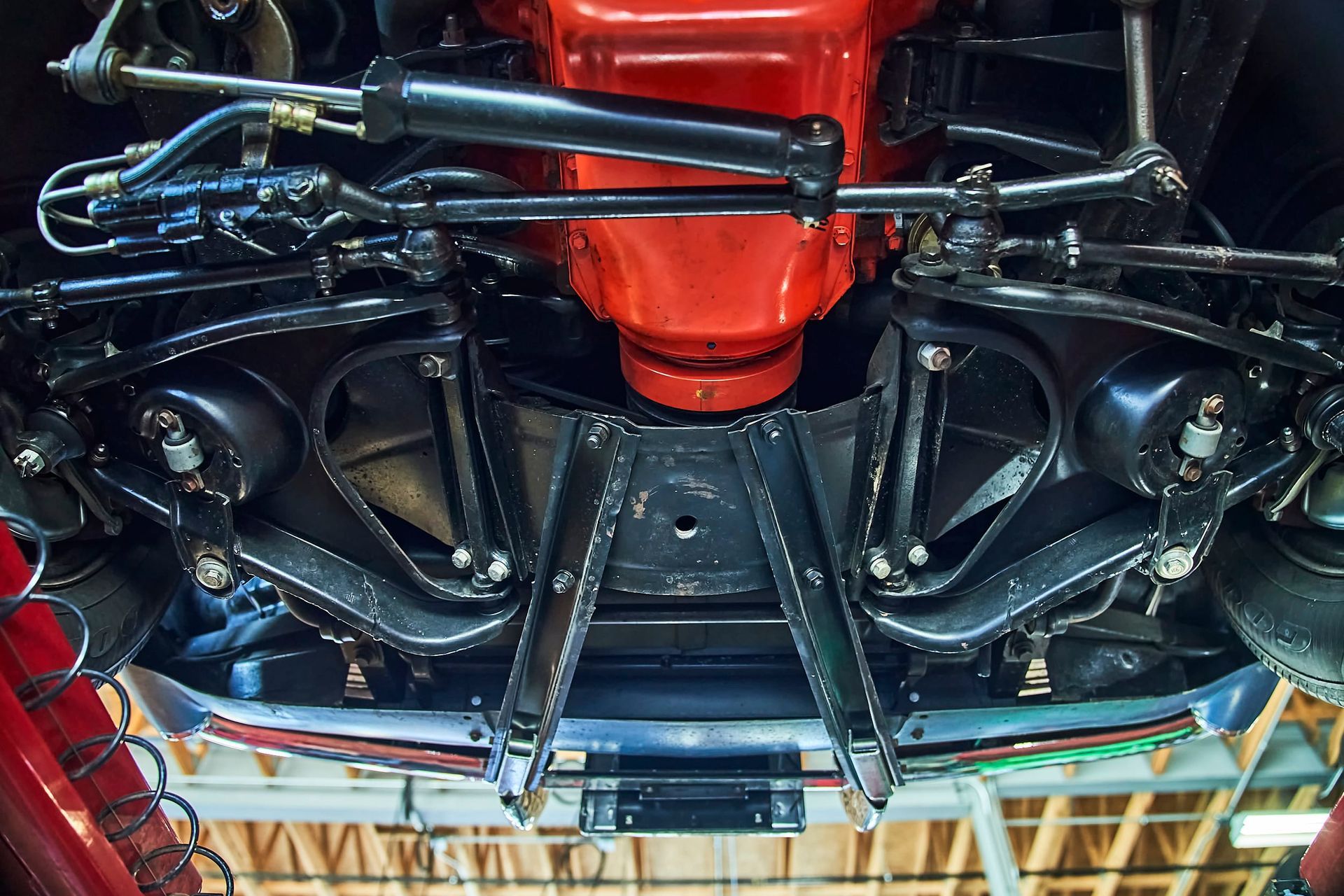 Underside of a classic car, showcasing the front suspension and red engine components. Dark metal contrasts with the bright engine color.