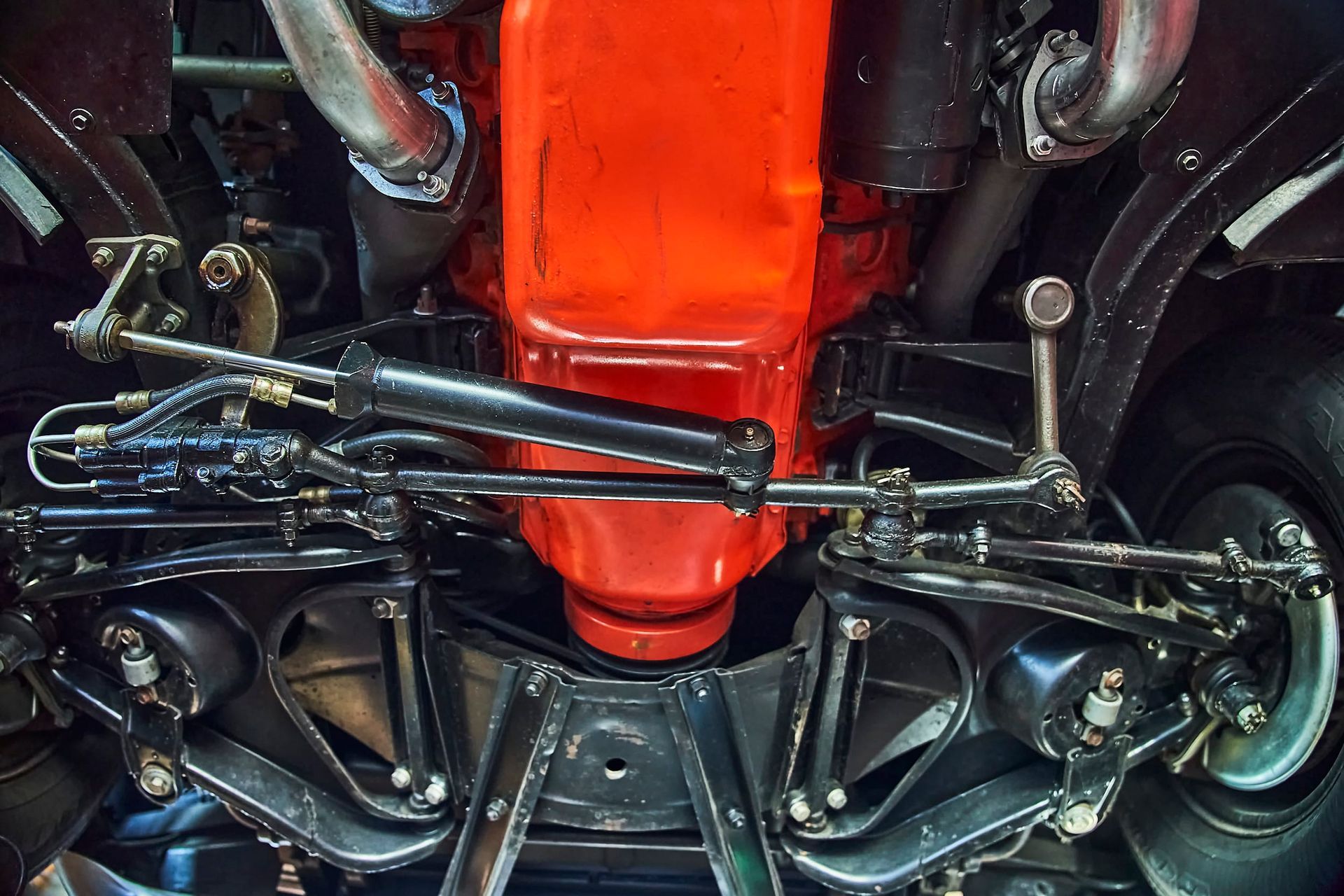 Underside view of a classic car engine painted bright red, surrounded by black chassis components and steering mechanisms.