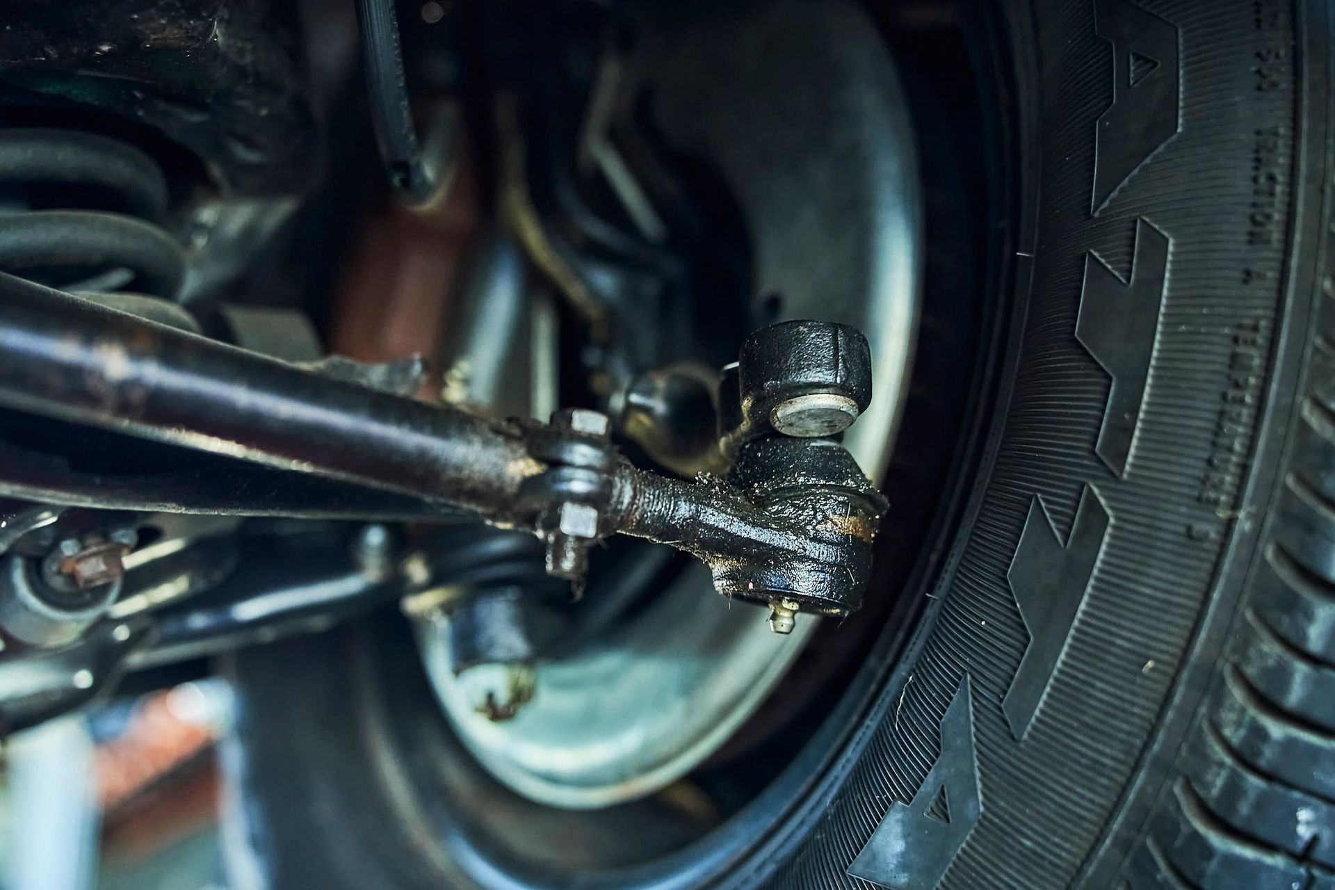 Close-up of a car's front suspension, focusing on the steering linkage and tire. The parts are black and greasy, alongside a black tire.