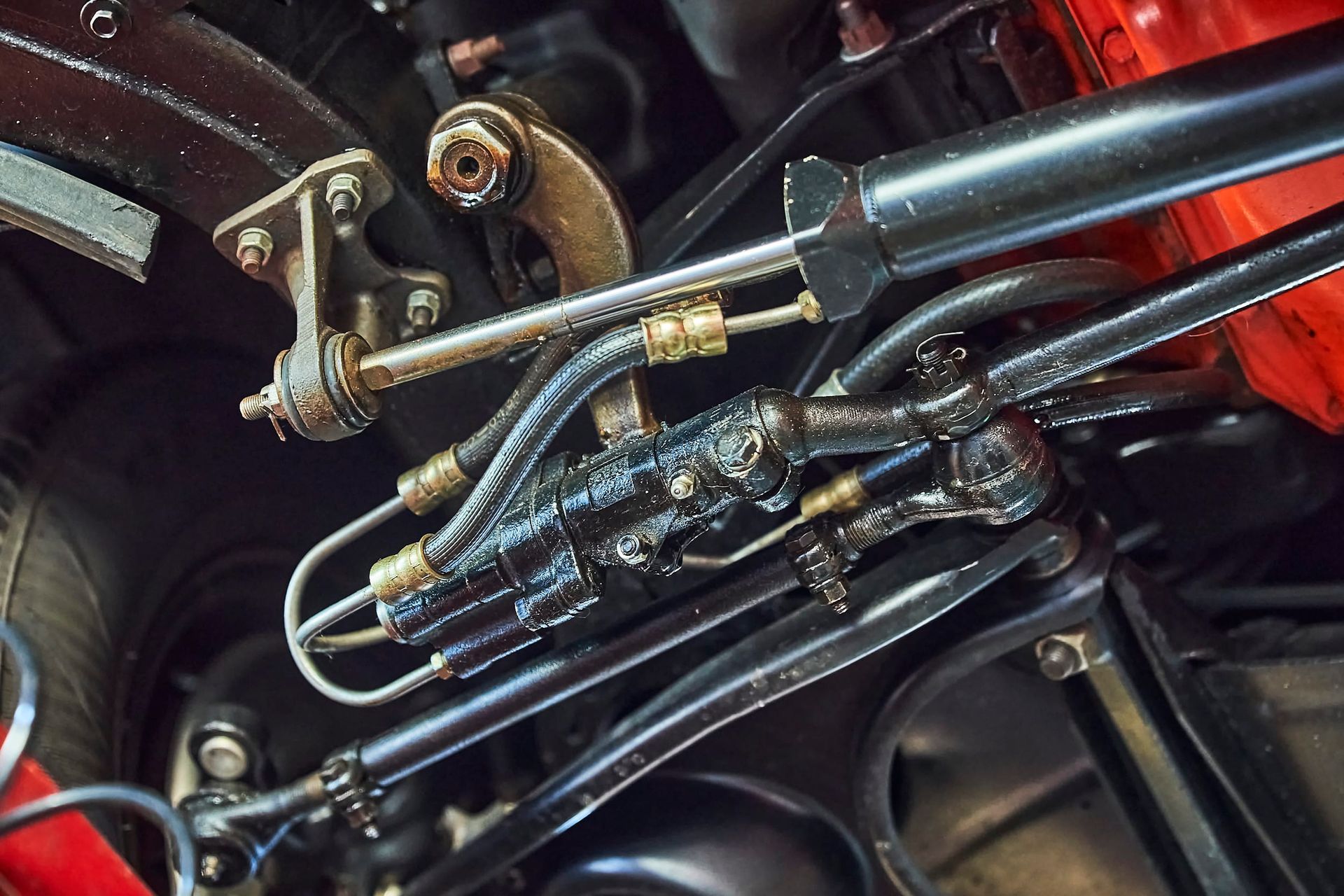 Close-up of car suspension components, featuring black steering parts, metal rods, and hoses. Red body panel visible.