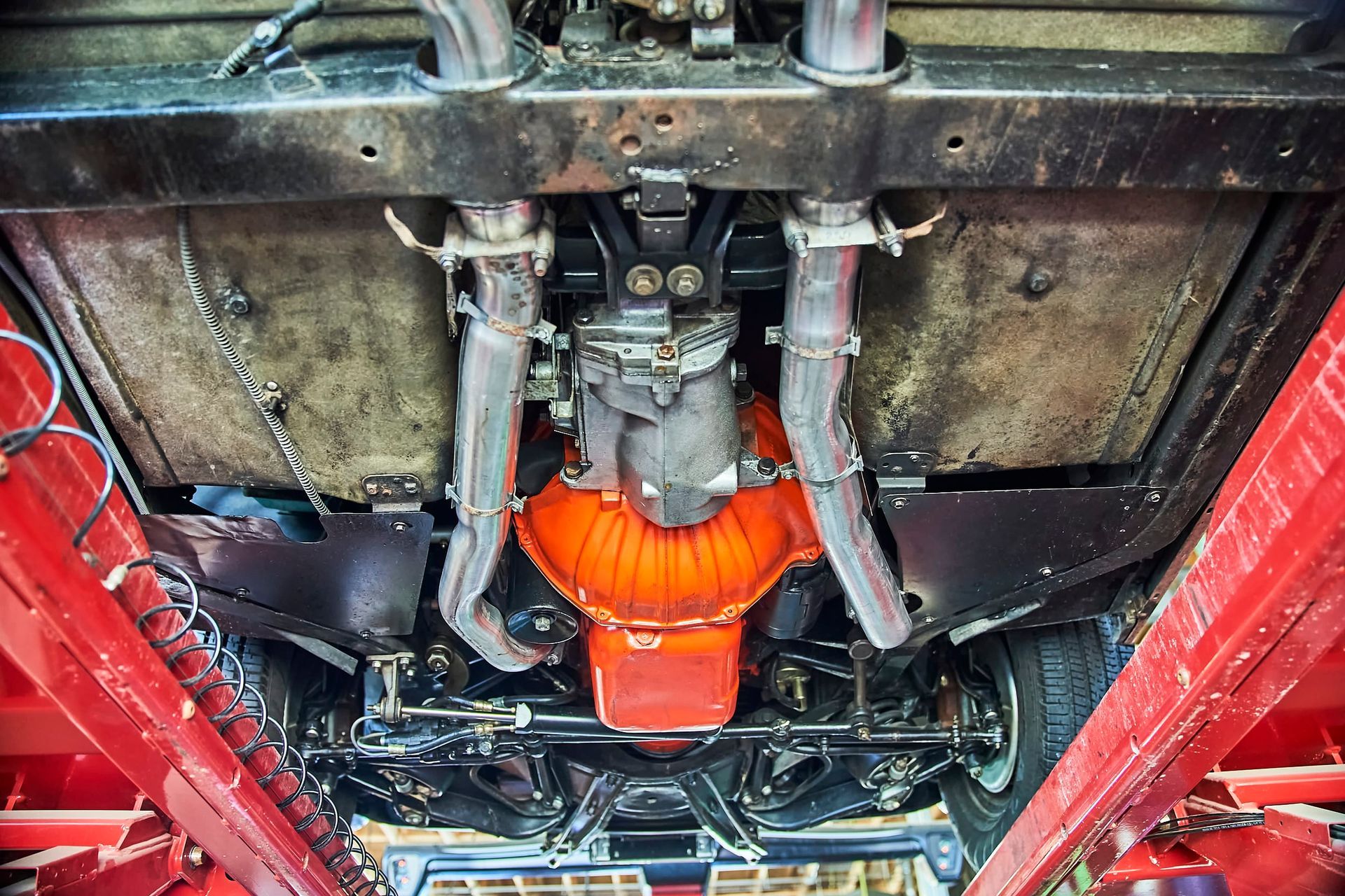Underside of a classic car, showing the orange engine, exhaust pipes, and undercarriage components. The vehicle is on a red lift.