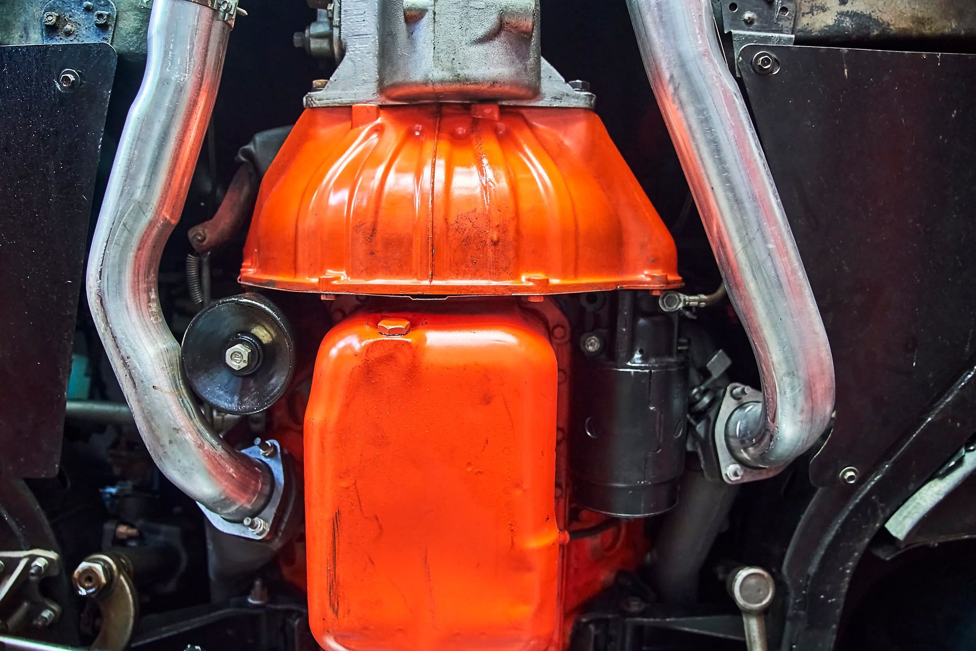 Orange engine components, including a transmission and oil pan, framed by chrome exhaust pipes.