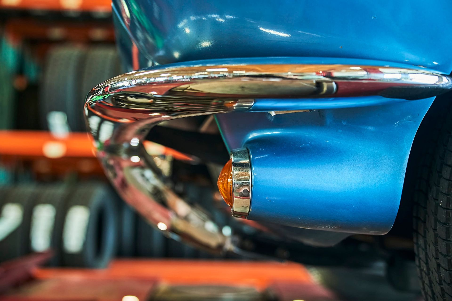 Close-up of a blue car bumper with chrome detailing and an orange side marker light, tires in the blurred background.