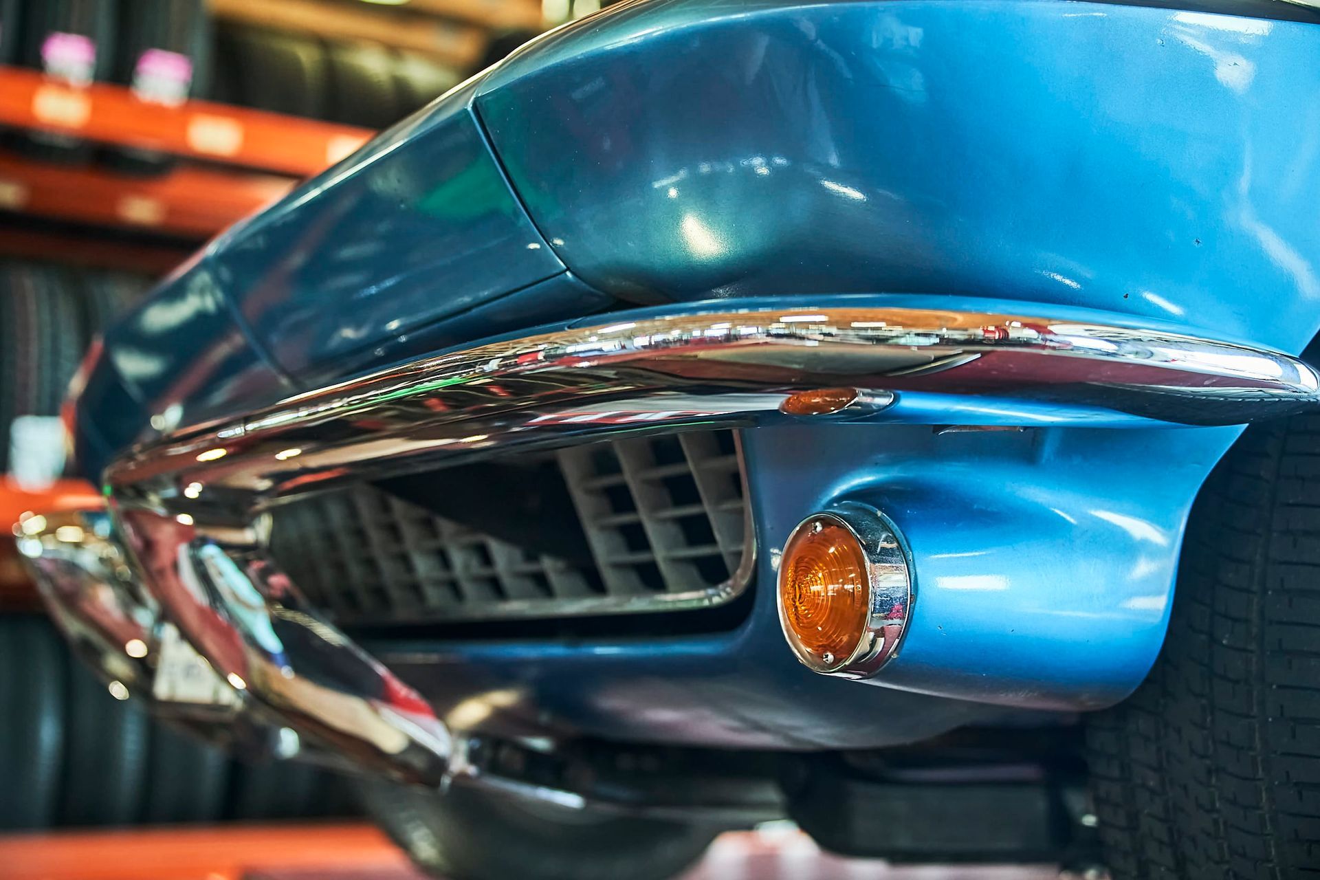 Close-up of a classic blue sports car's front end with chrome accents, including the bumper, grill, and amber turn signal. Inside a garage with tires on shelves.