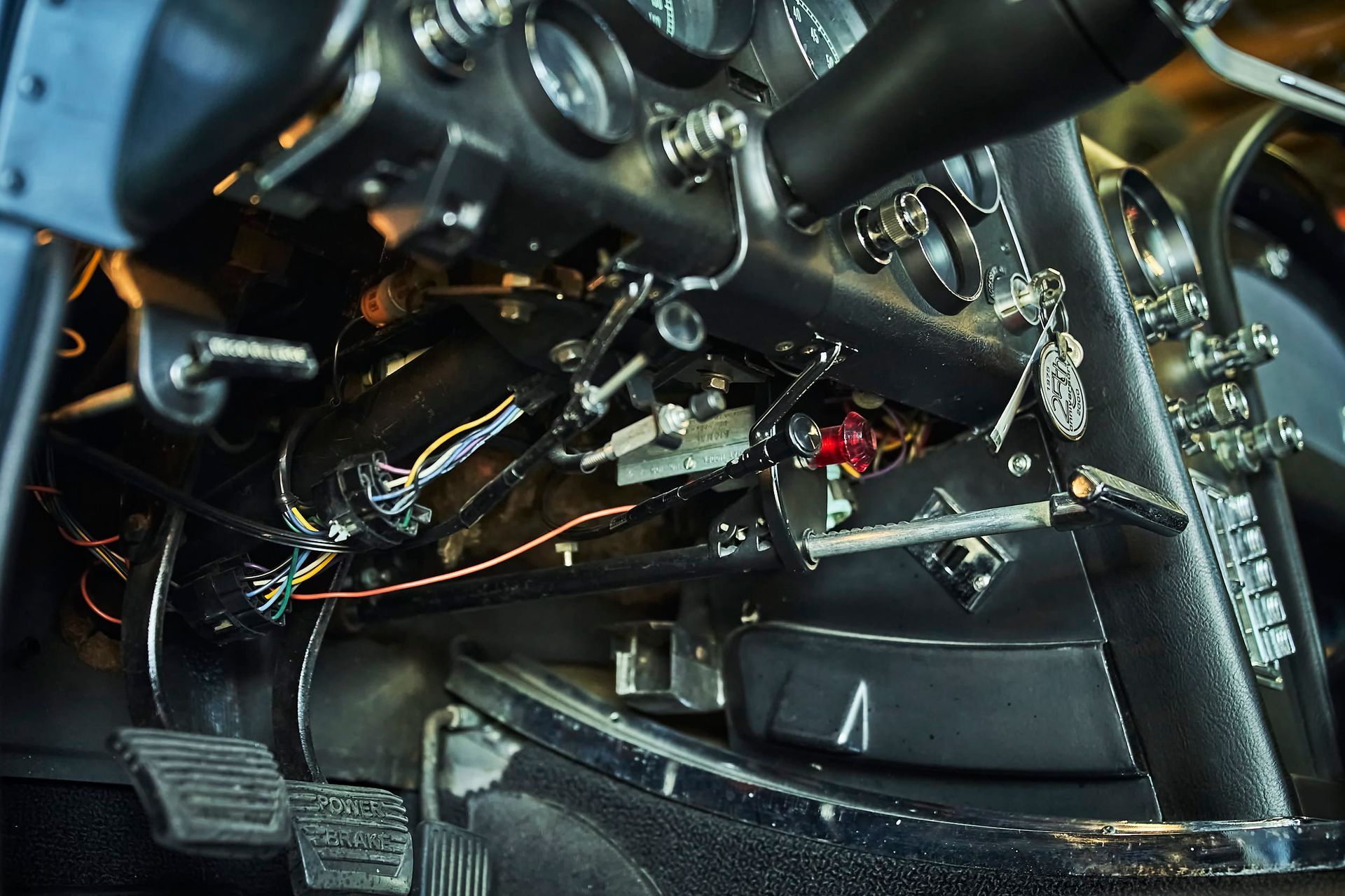 Dashboard and pedals of a classic car with exposed wiring and gauges visible. The interior is dark with various levers and details.