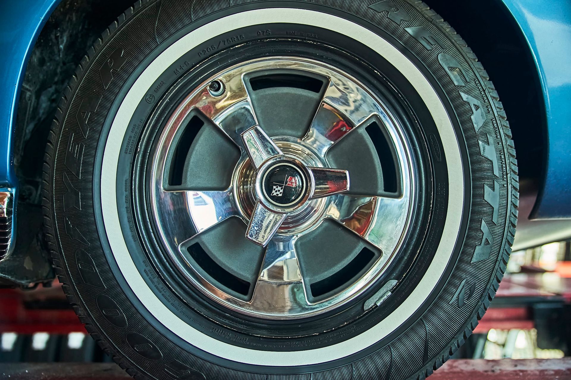 Close-up of a blue car wheel with a chrome hubcap and white sidewall tire. The wheel is set in a garage setting.