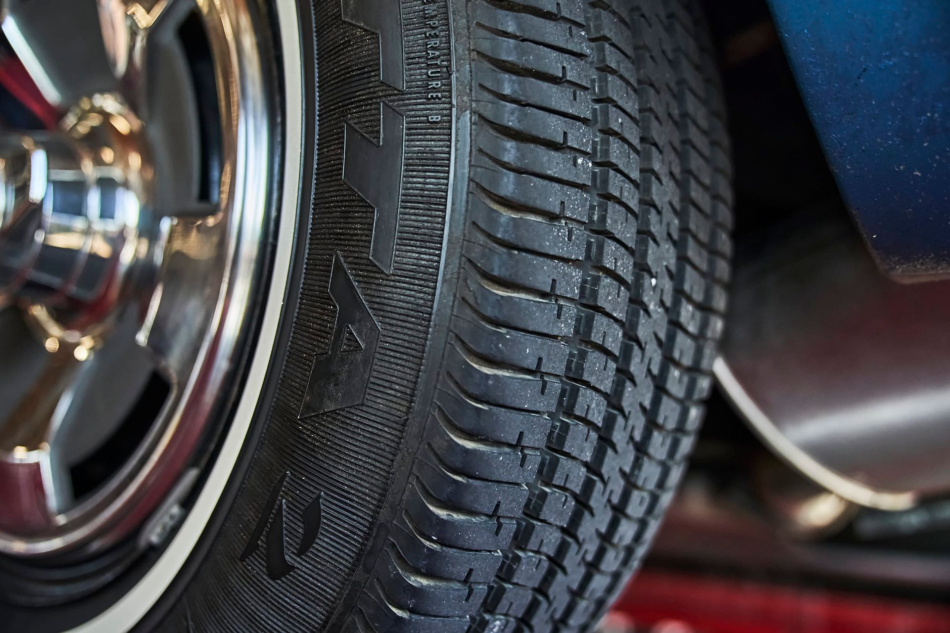 Close-up of a car tire with a white sidewall. The tire has a black tread pattern with a shiny chrome rim and a blue car body is visible.