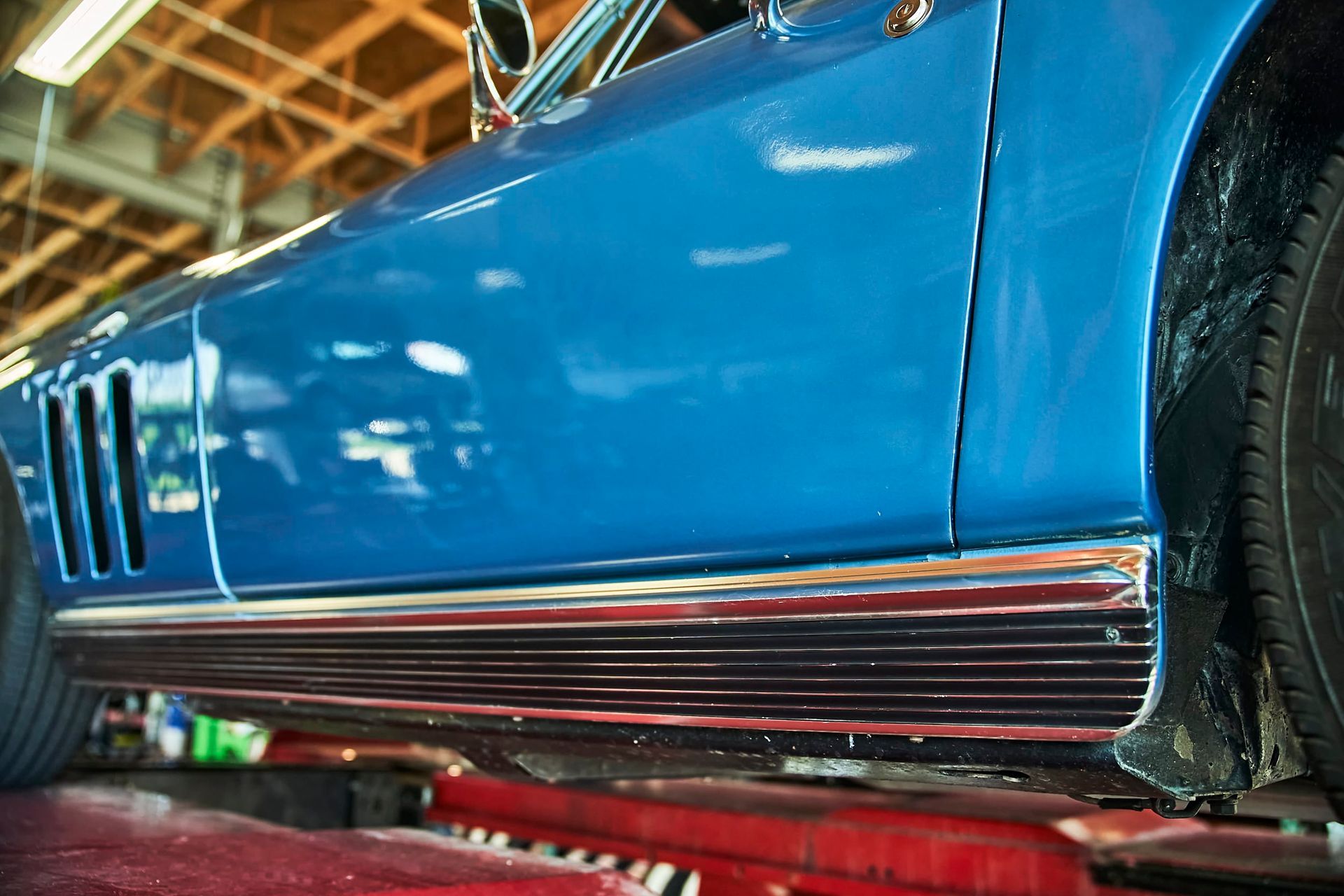 Blue classic car's side, showing chrome trim, tire, and vents, likely in a repair shop.