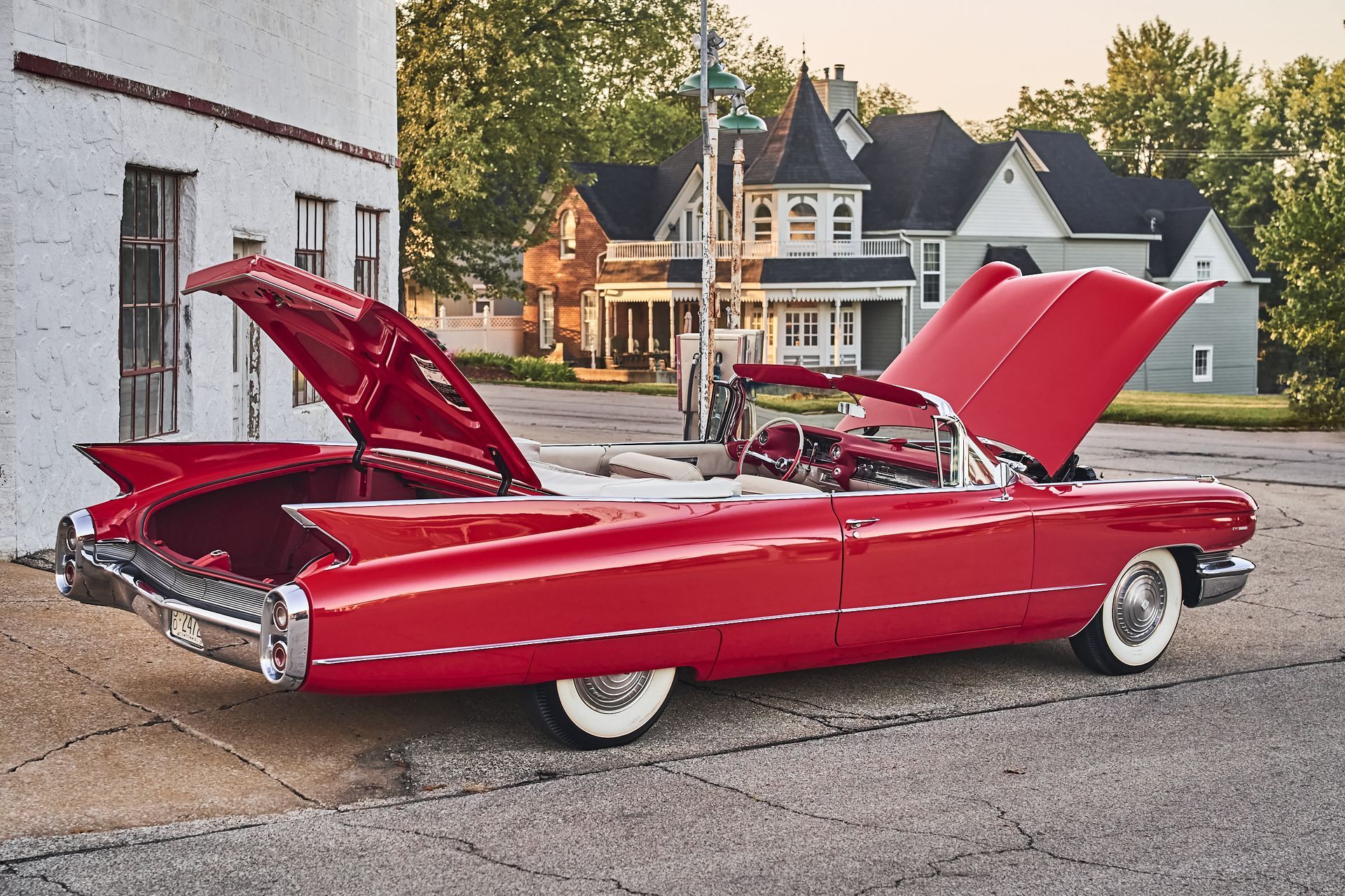 A red cadillac with the hood up is parked in front of a white building.