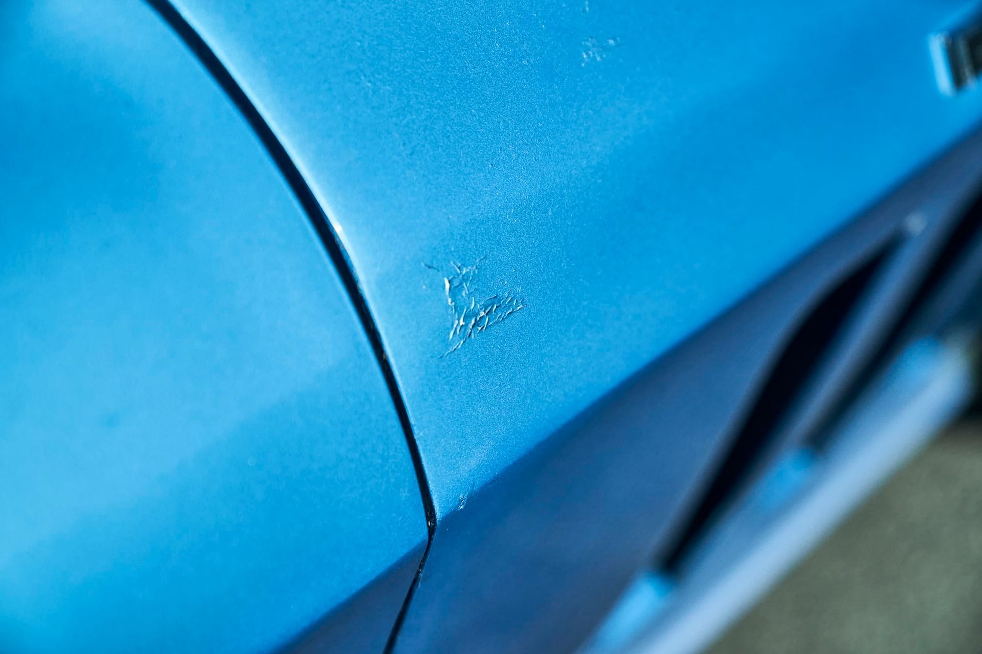 Close-up of a blue car's body with noticeable paint chips. The damage is located near a body seam.