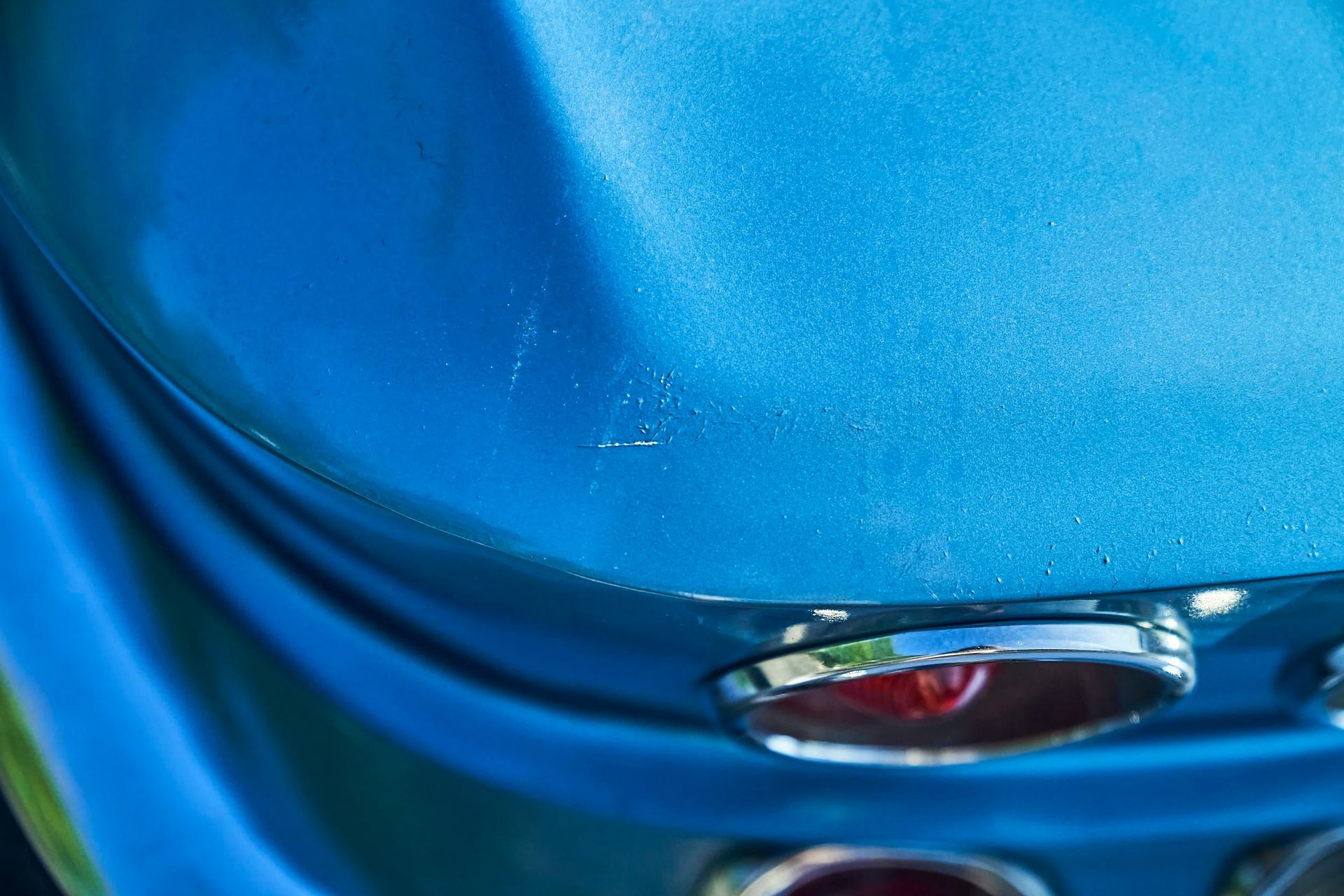 Close-up of a blue car's rounded back end.  Small paint chips are visible near the top, above a chrome-ringed taillight.