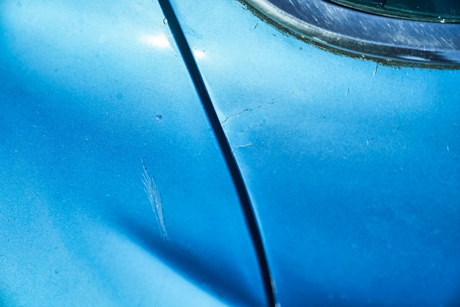 Close-up of a blue car's body with scratches and a dark vertical seam near a window.