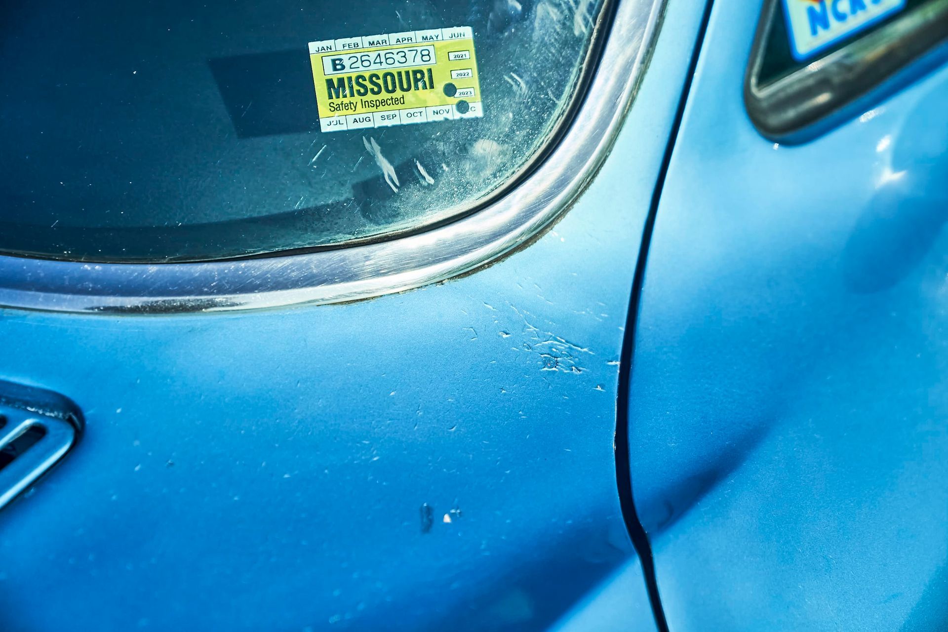Close-up of a blue car, showing a Missouri license plate sticker on the windshield and part of the car's side.