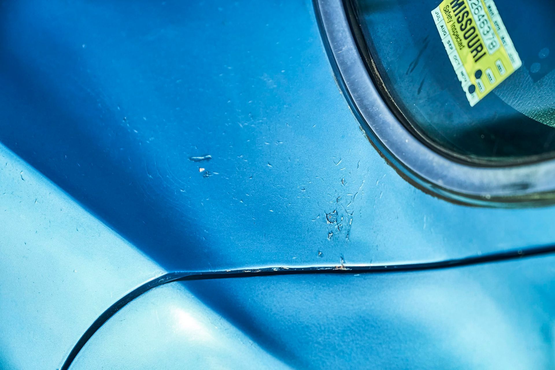 Close-up of a blue car's body, showing paint damage and a Missouri license plate sticker near the window.