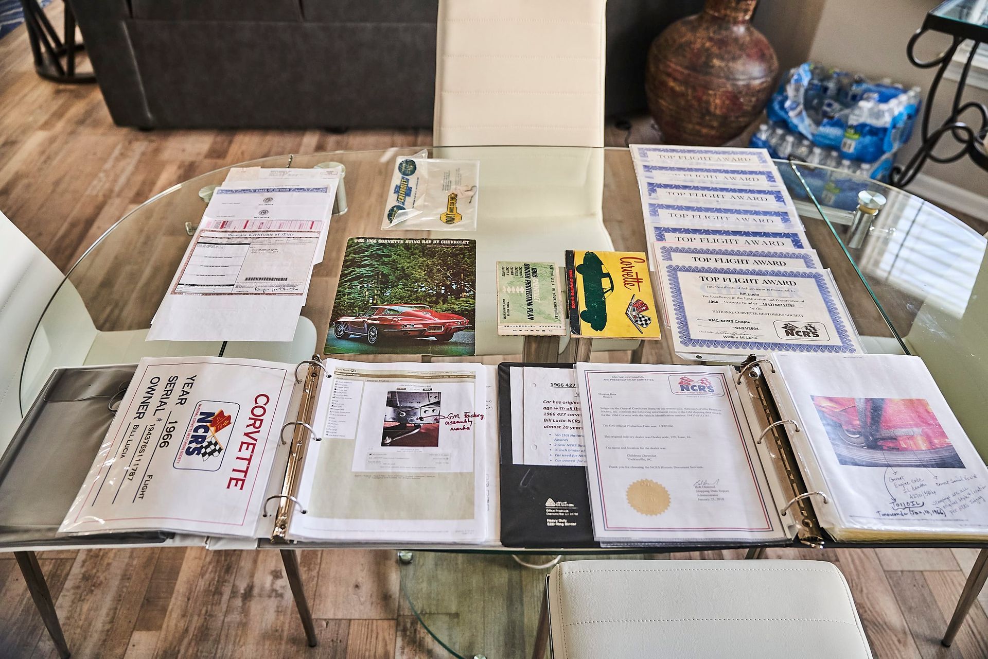 Papers and documents spread on a glass table, including binders and certificates, suggesting car documentation.