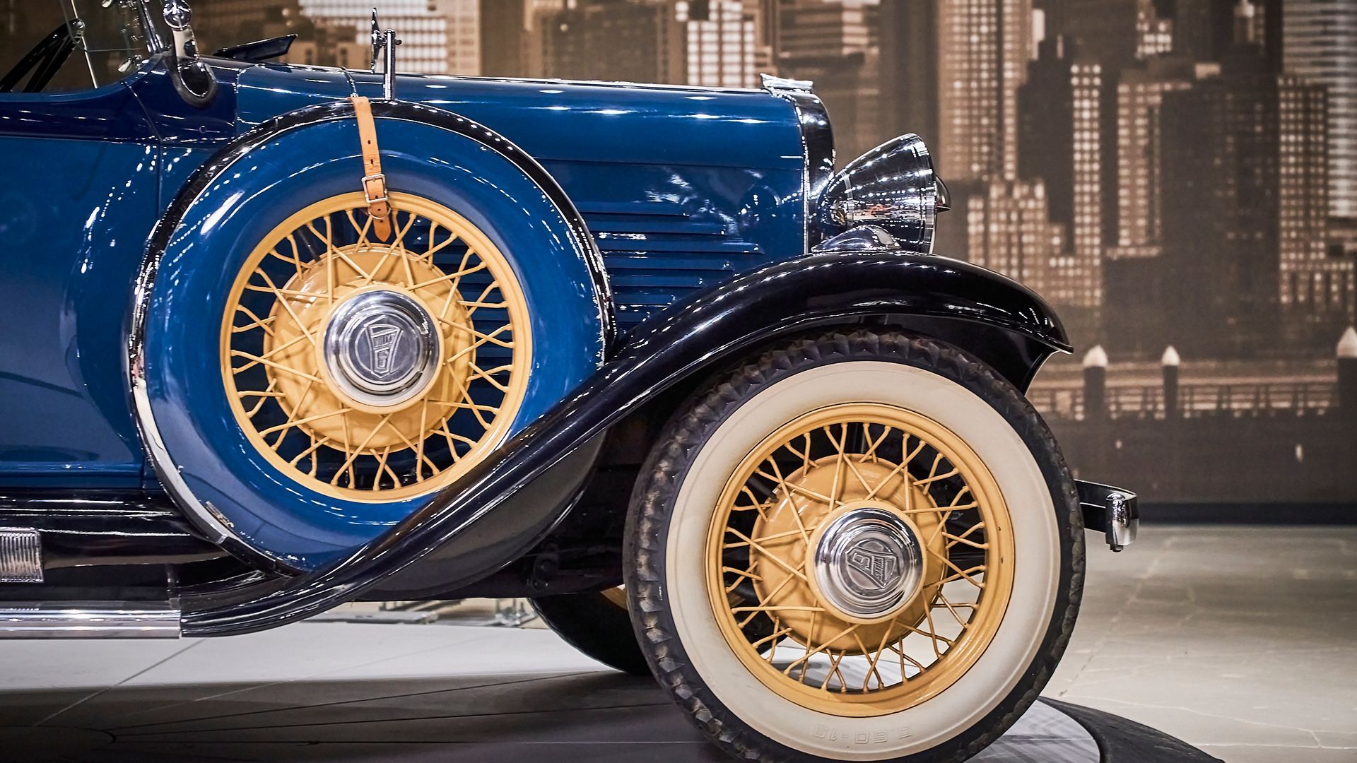 A blue car with gold rims and white tires is parked in front of a city skyline.