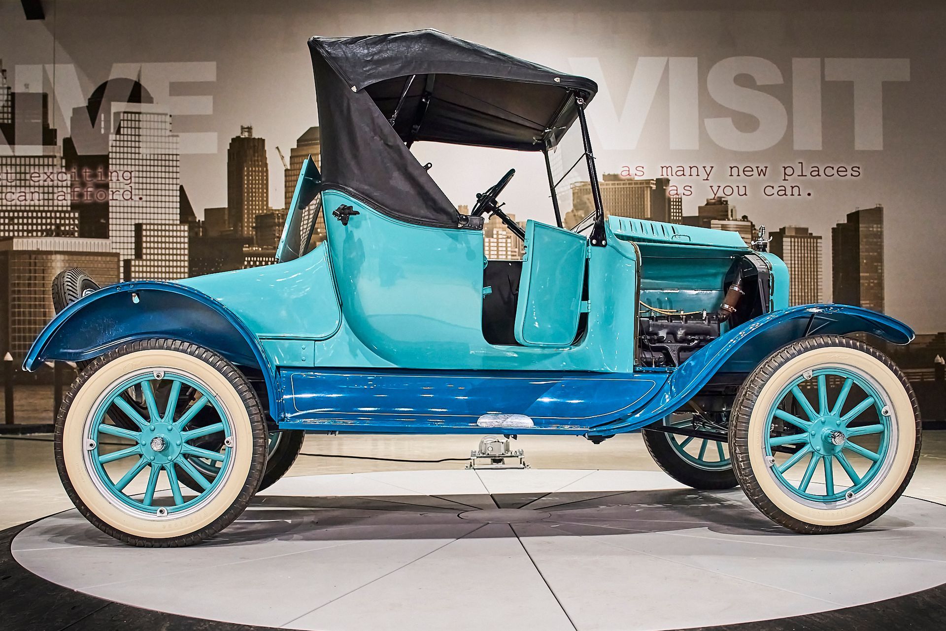 A blue car with white tires is on display in front of a sign that says visit