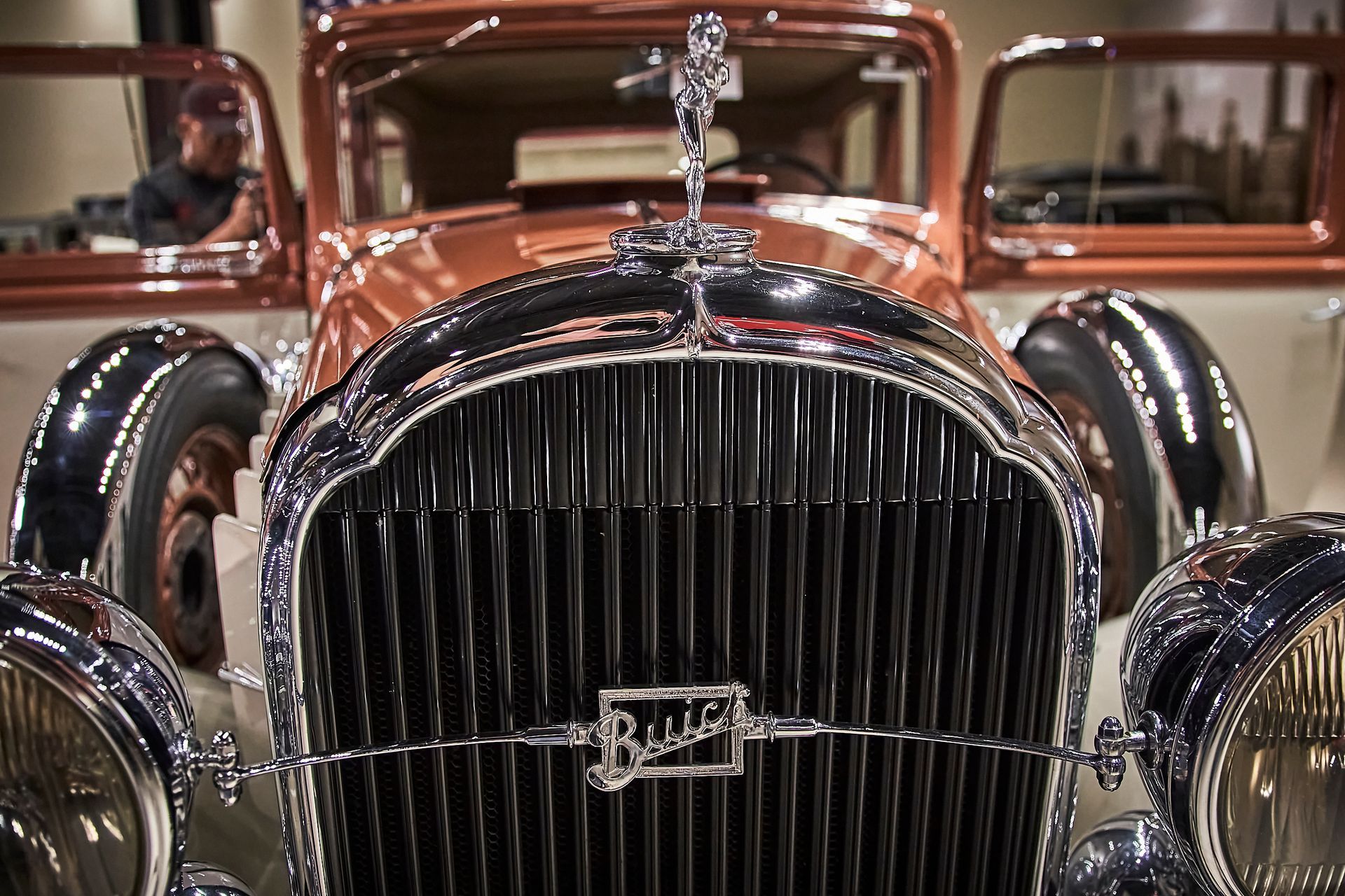 A close up of the front of an old car with a man driving in the background.