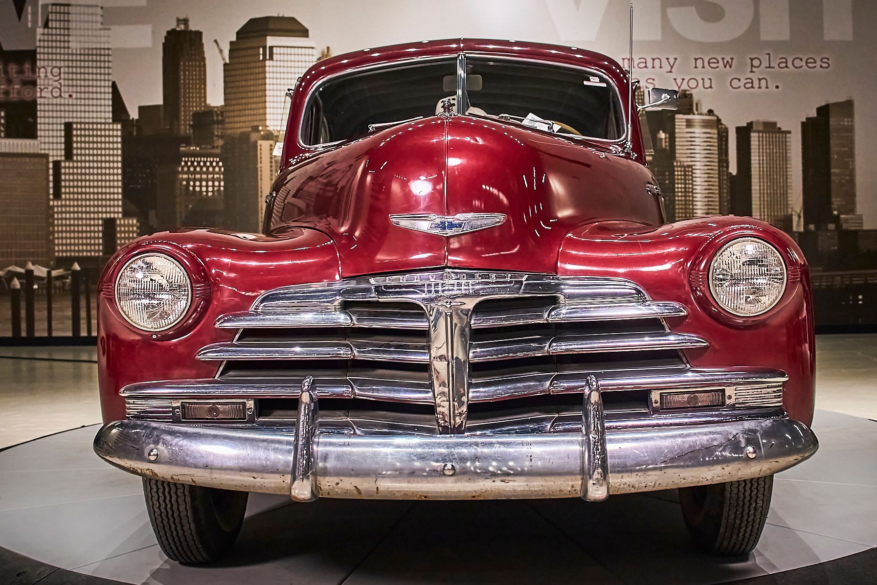 An old red car is parked in front of a city skyline