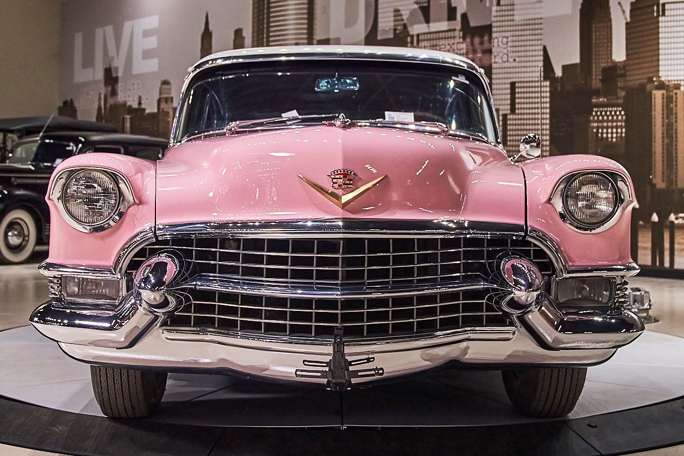 A pink cadillac is parked in front of a wall that says live