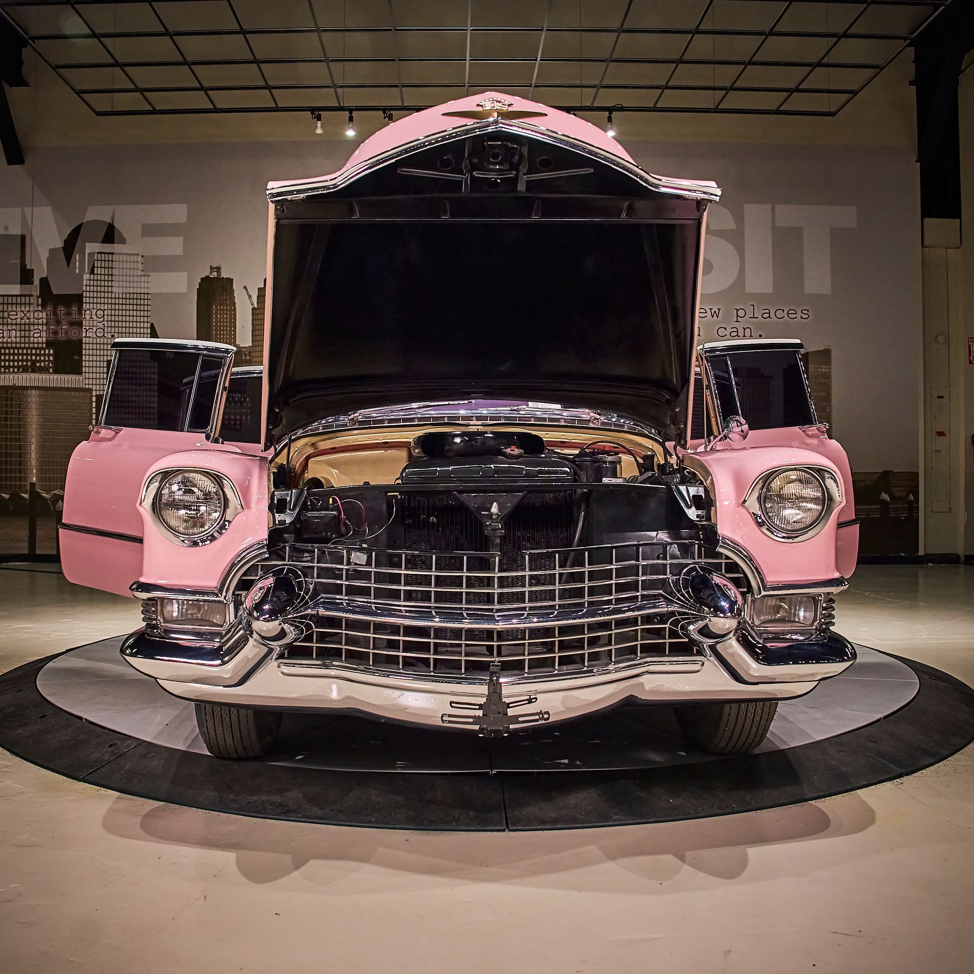A pink car with the hood open is sitting in front of a sign that says we sit