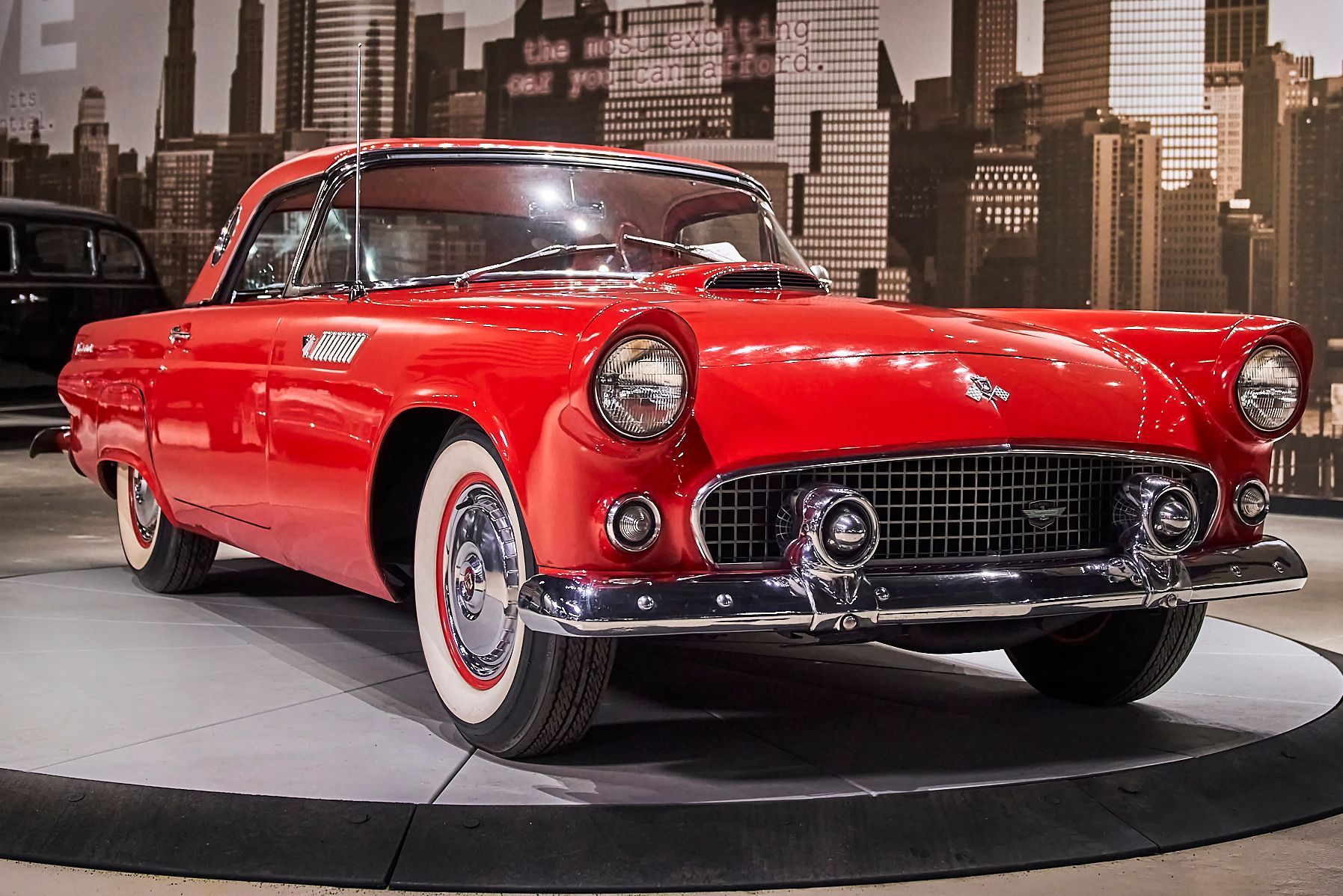 A red car is on display in front of a city skyline.