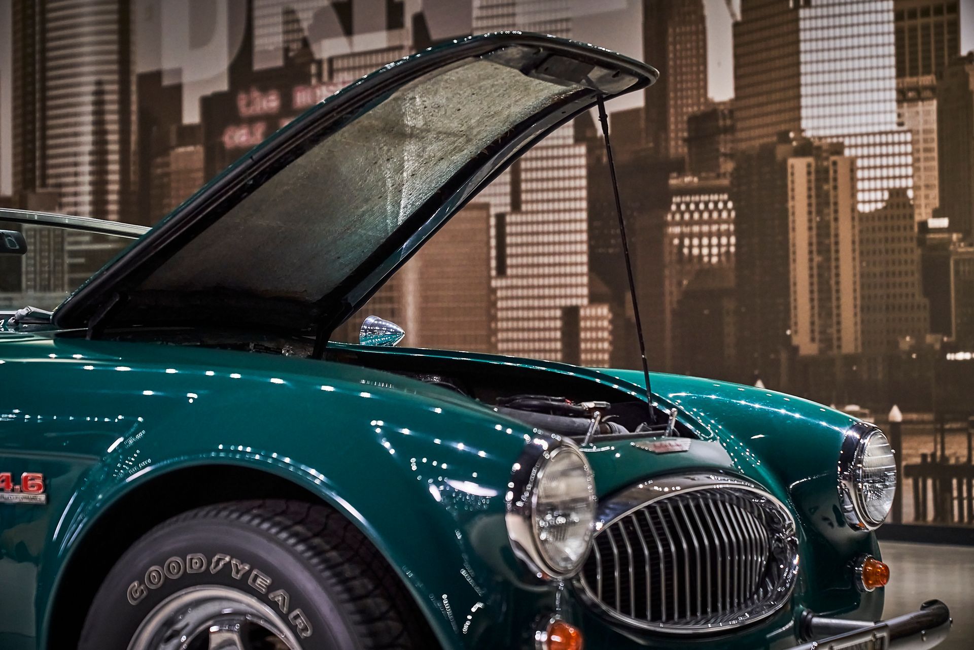 A green car with the hood up is parked in front of a city skyline.