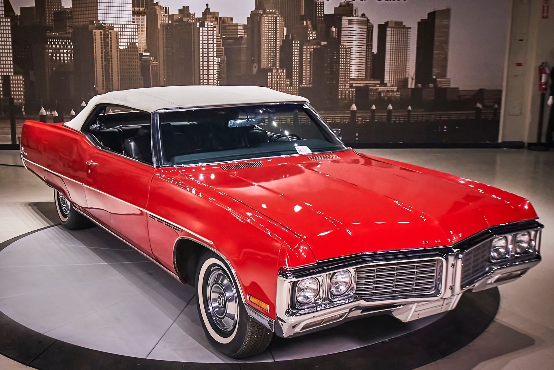 A red car with a white top is parked in front of a city skyline