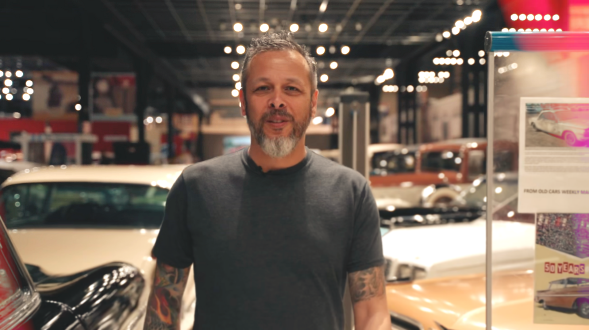 A man with a beard and tattoos is standing in front of a car.