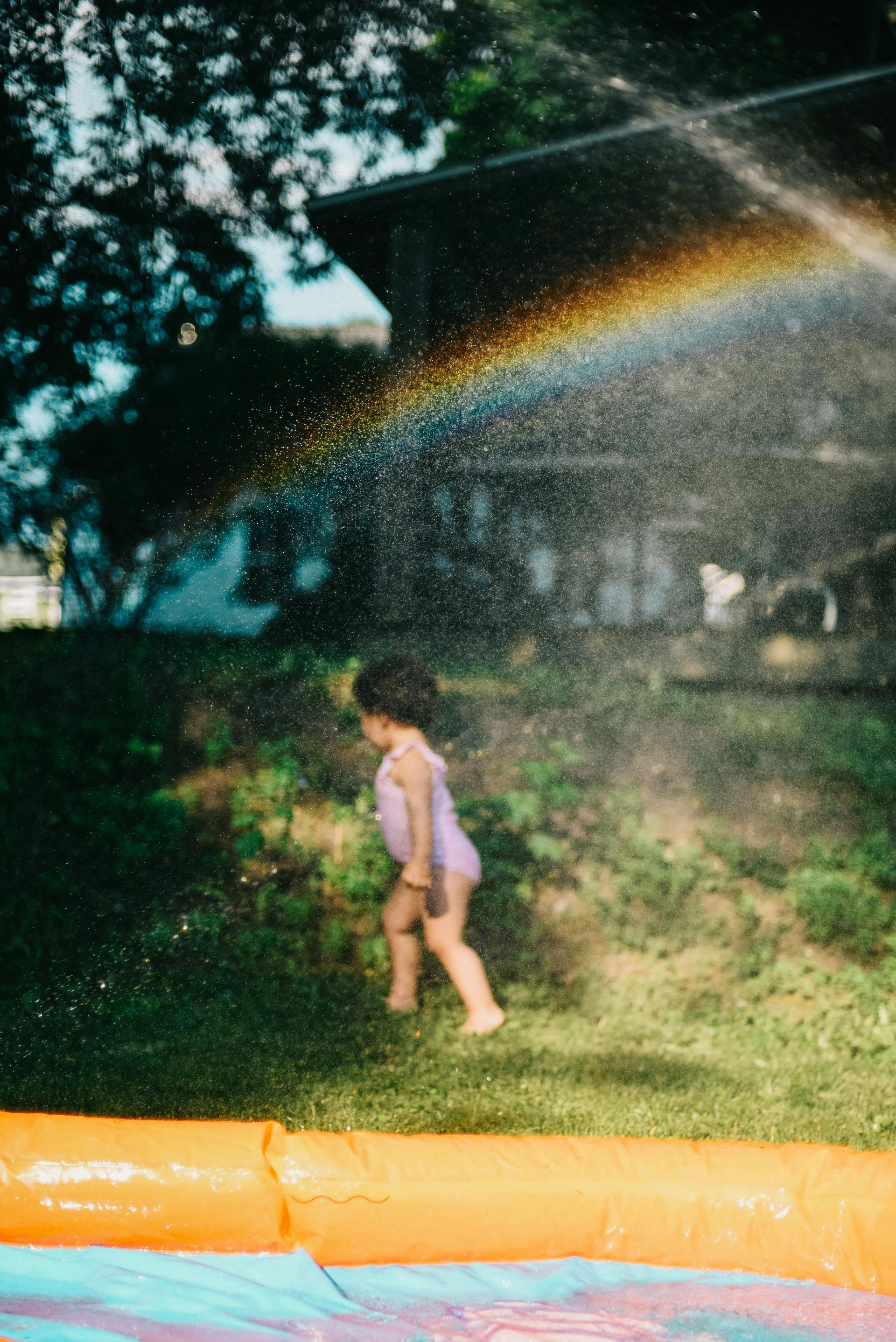 Een klein meisje speelt op een waterglijbaan met een regenboog op de achtergrond.