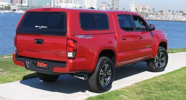 Red Toyota Tacoma pickup truck with a camper shell parked by the water.