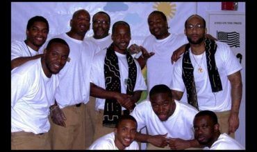 Group of men in white shirts, some with black scarves, posing indoors.